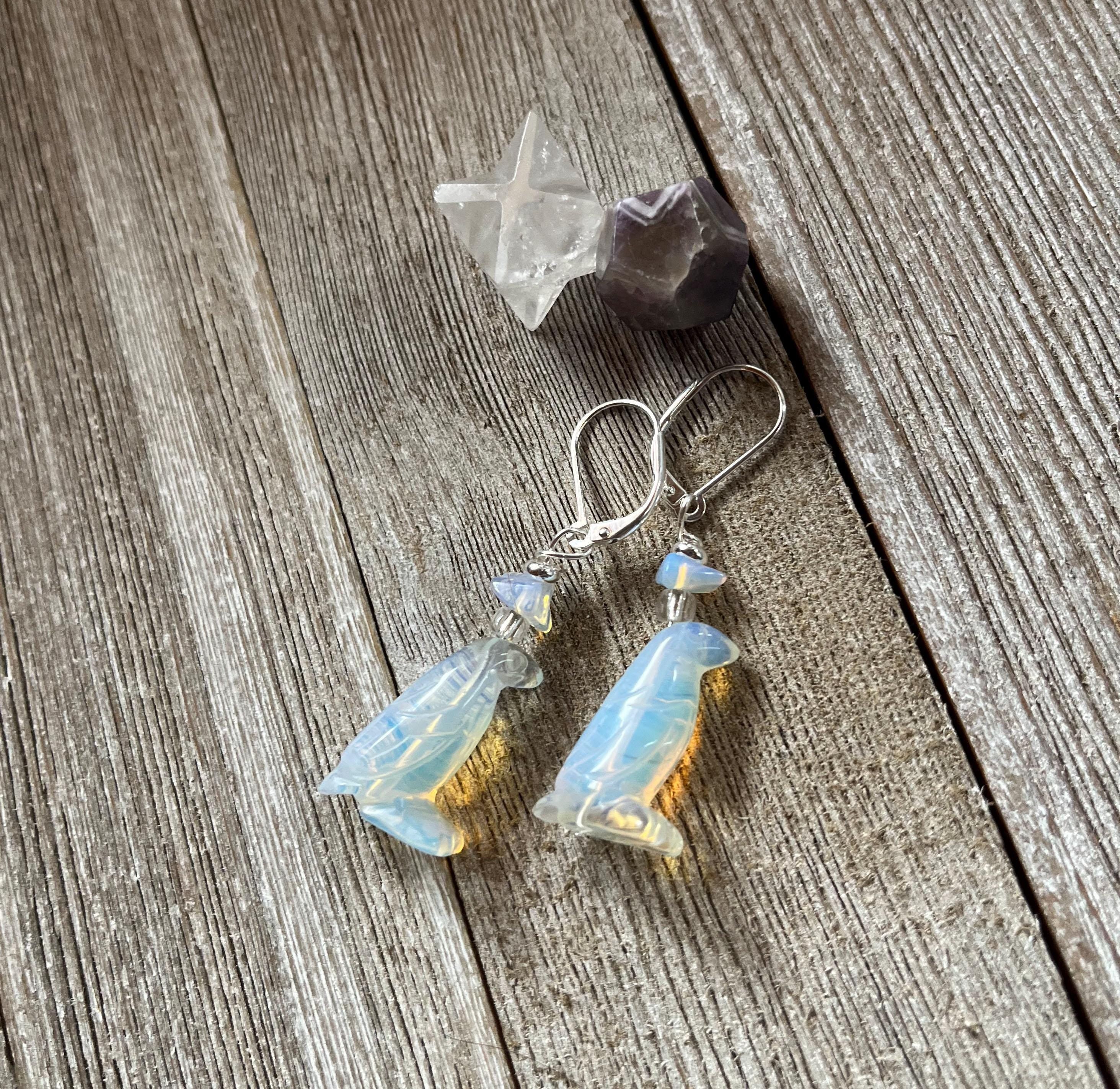 Handcrafted Opalite Penguin Earrings with sterling silver lever backs rest on rustic wooden planks, accompanied by a clear star-shaped crystal and a faceted purple stone.