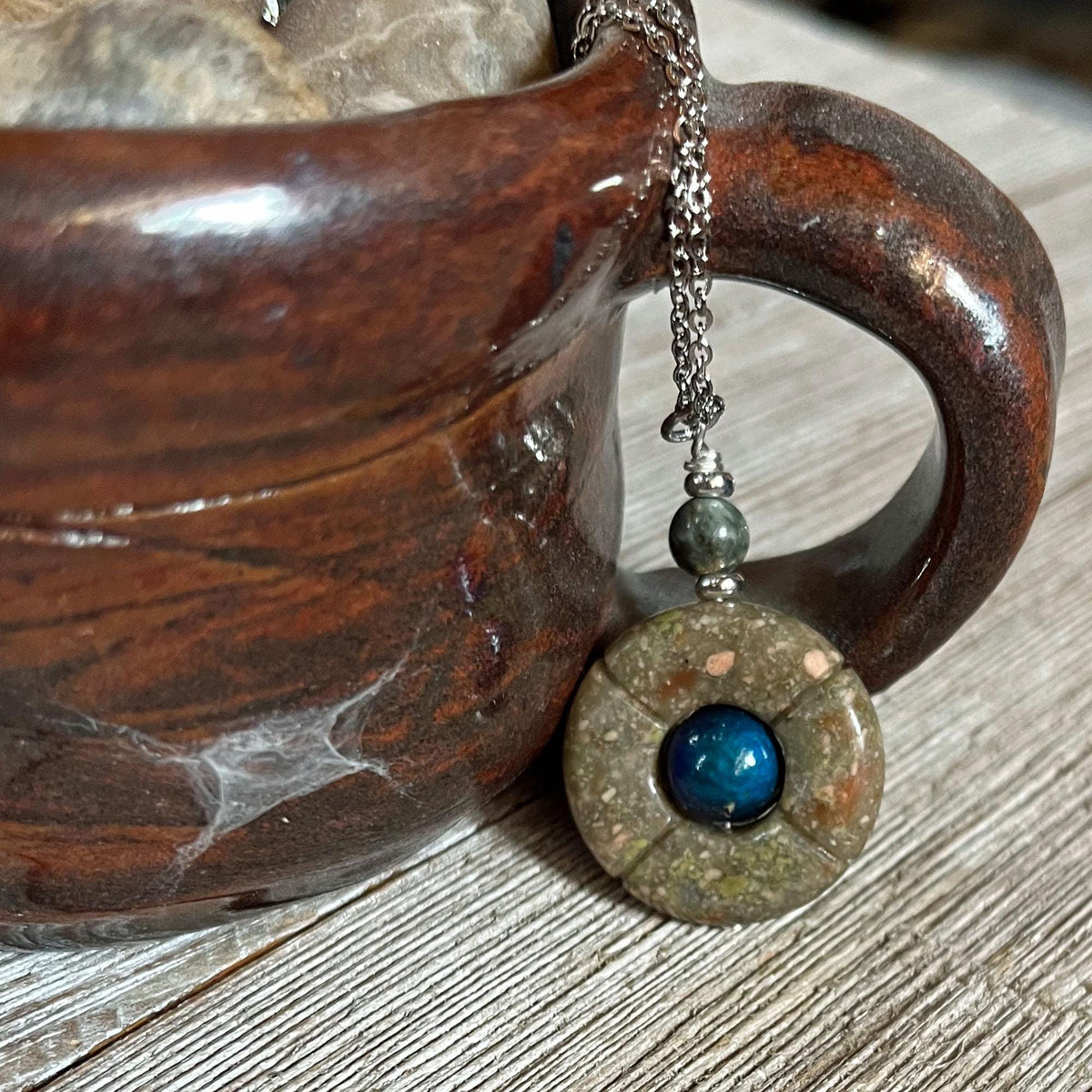 The One of a Kind Jasper Mood Pendant, featuring a color-changing bead on an 18” silver chain, hangs from the handle of a brown ceramic mug resting on a wooden surface.