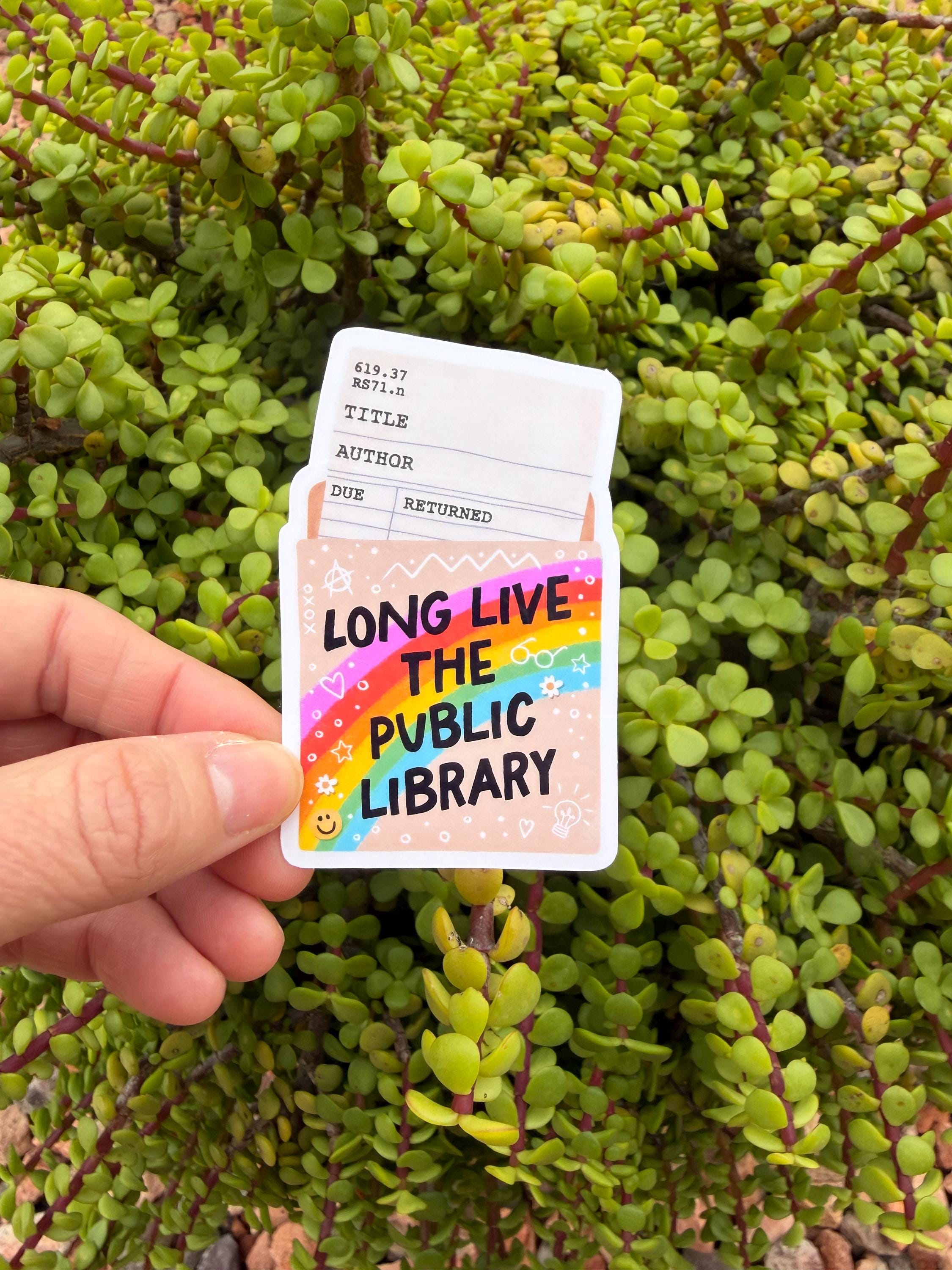 May include: A sticker with a library card design and the text "LONG LIVE THE PUBLIC LIBRARY." The sticker features a rainbow, stars, and hearts. The background is a green plant.