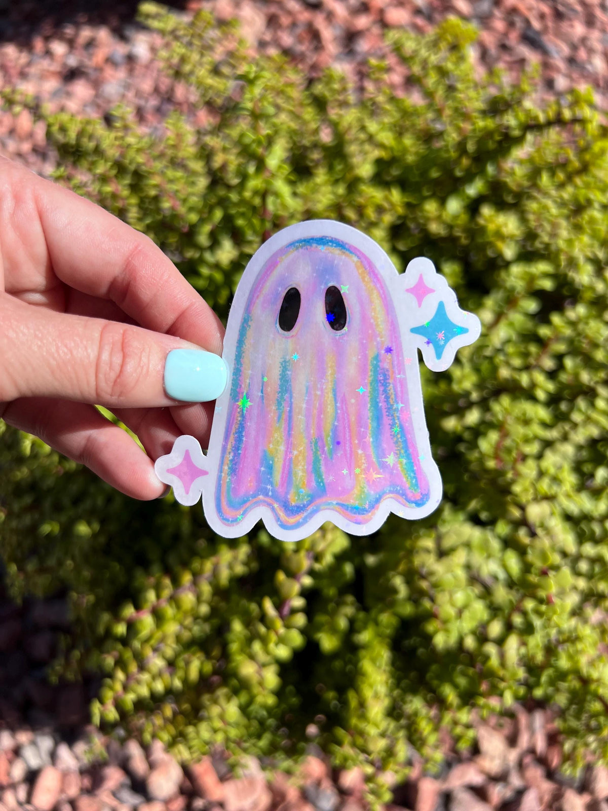 May include: A colorful ghost sticker with a rainbow design and glitter accents. The sticker has a white border and is decorated with two black eyes and two starbursts. The sticker is held in front of a green bush.