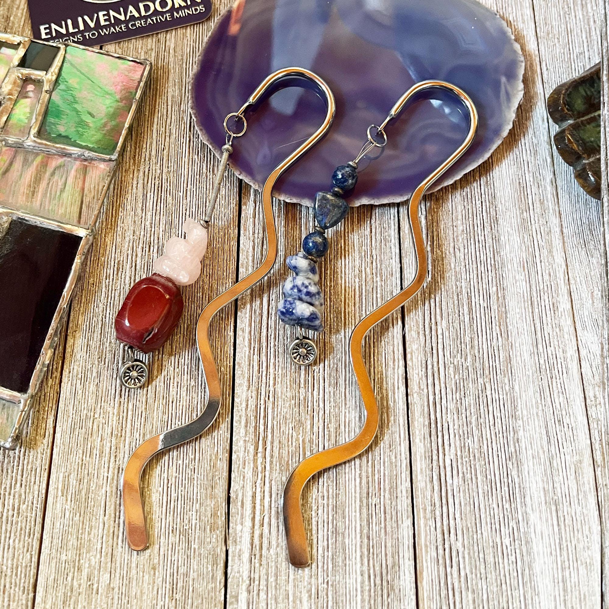 Two Snake Bookmarks—Rose Quartz Serpent and Blue Sodalite—from the Reptile Reader Gift set are displayed on a light wood surface with stained glass and a purple agate slice.