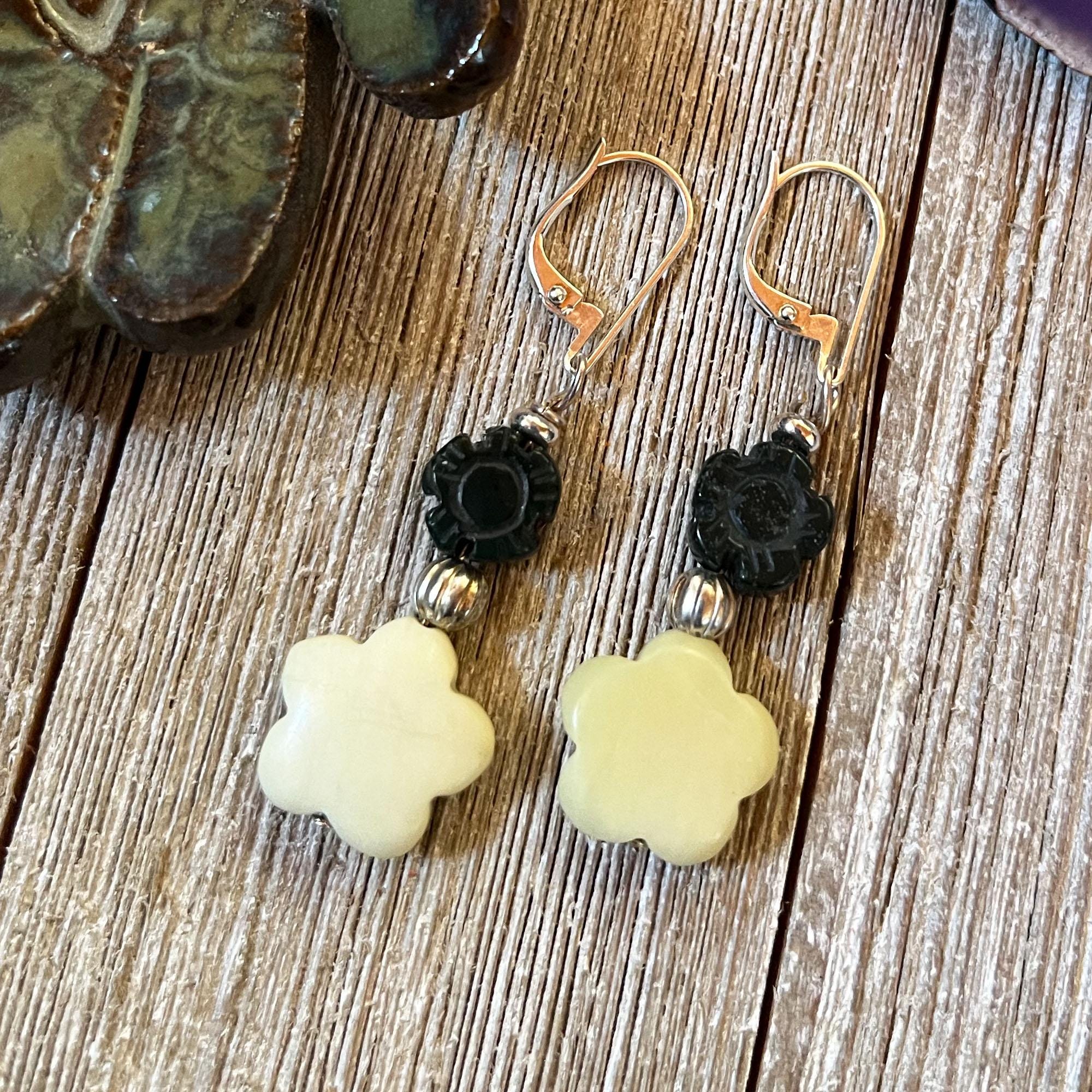 Spring Flower Earrings in carved stones, featuring cream flower-shaped beads, black carved beads, and silver spacers, perfect to complete your wardrobe and add cheerful style to daily wear. Displayed on a wooden surface by a ceramic object.