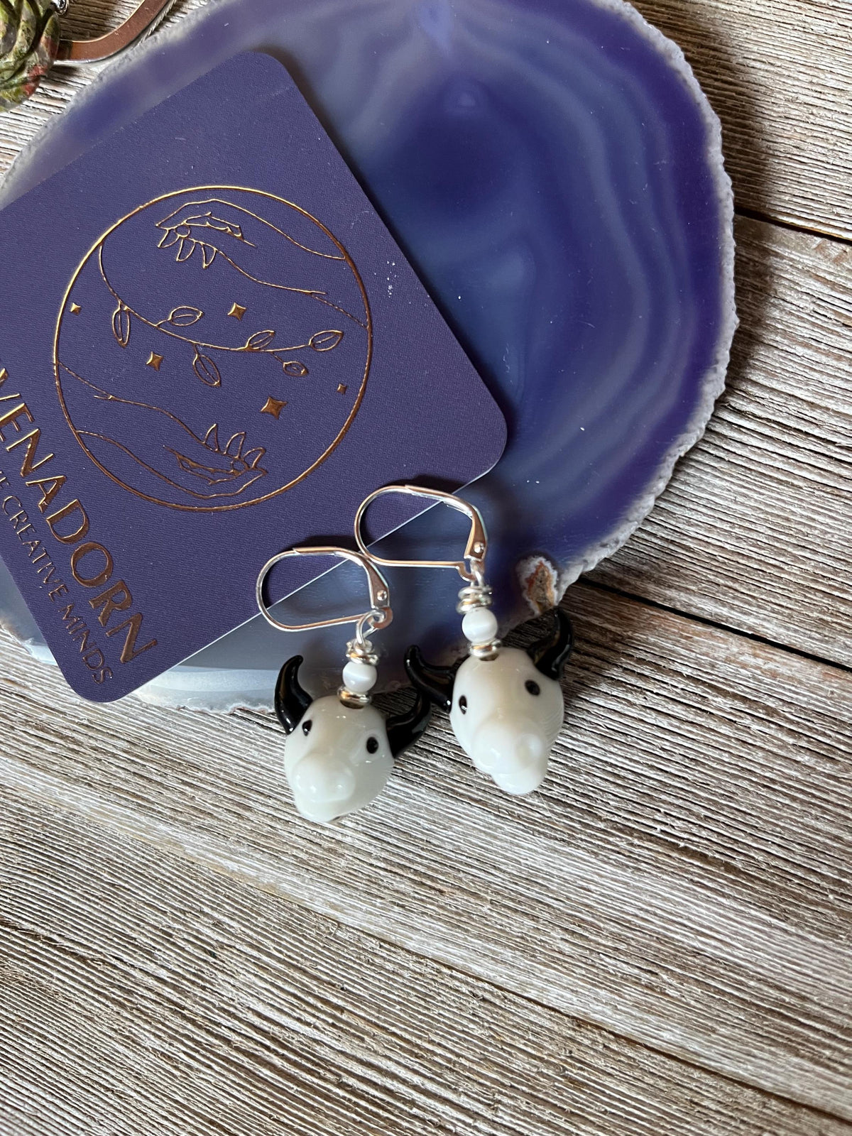 Handcrafted Glass Bull Head Earrings, featuring small white charms and pearls, displayed on a purple agate slice beside a card with gold celestial designs and LINADORN HANDCRAFTED JEWELS—an ideal animal lover jewelry gift.