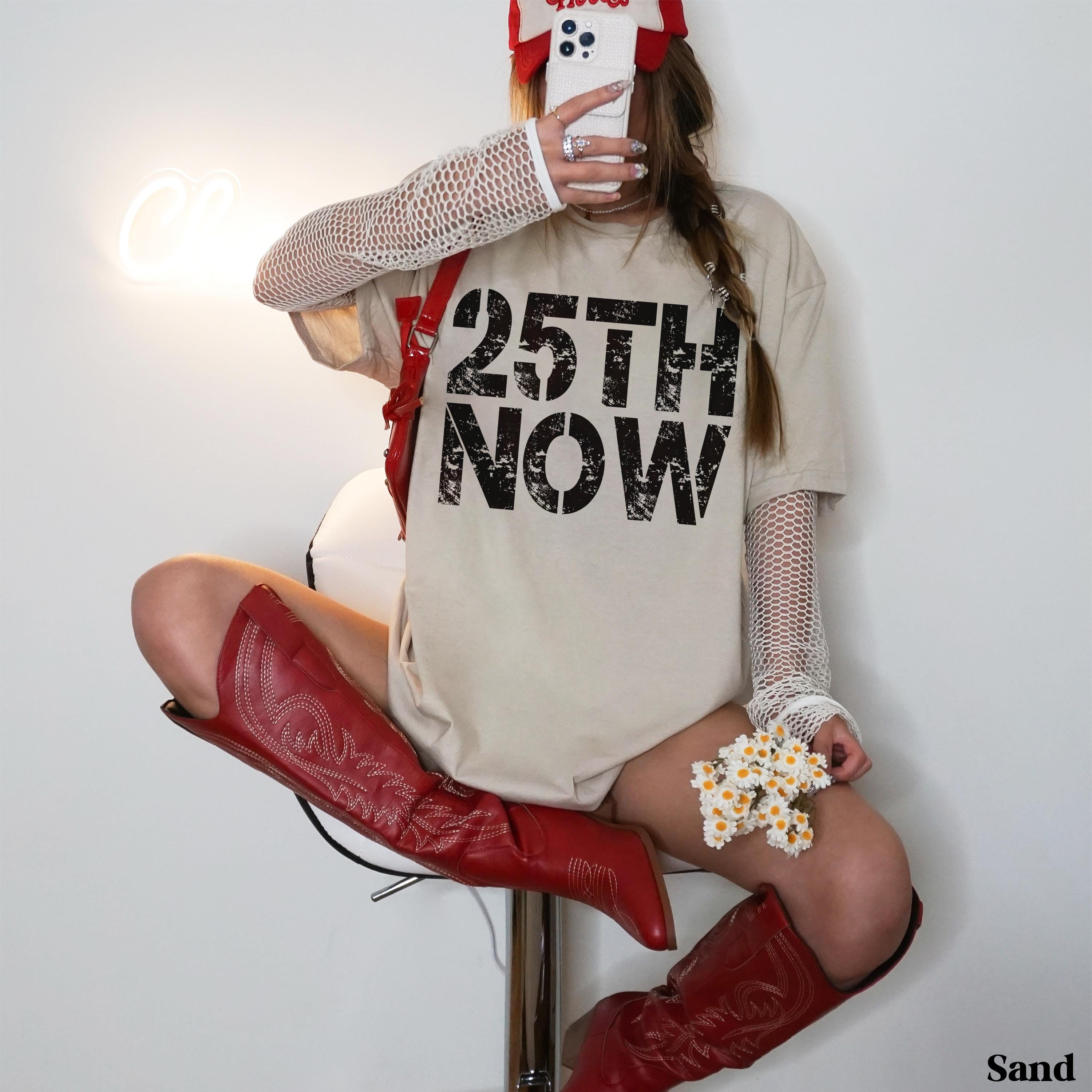 Woman in tan t-shirt with "25TH NOW" and red boots sits on stool.
