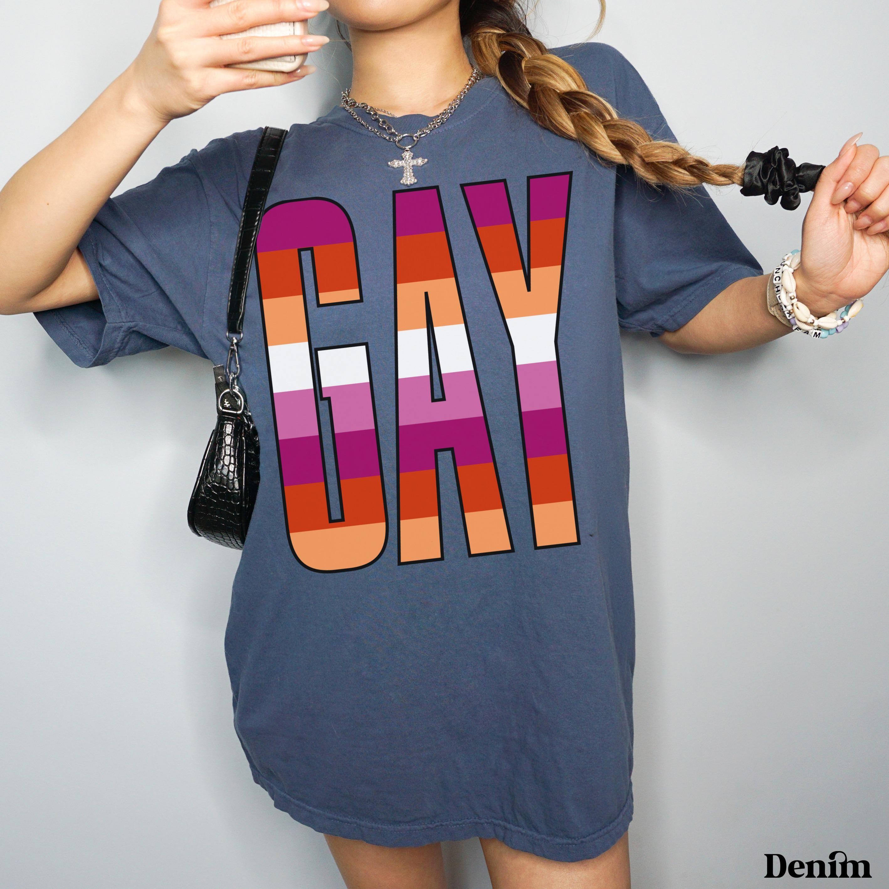 Woman in charcoal t-shirt with "GAY" in rainbow stripes, holding purse and phone.