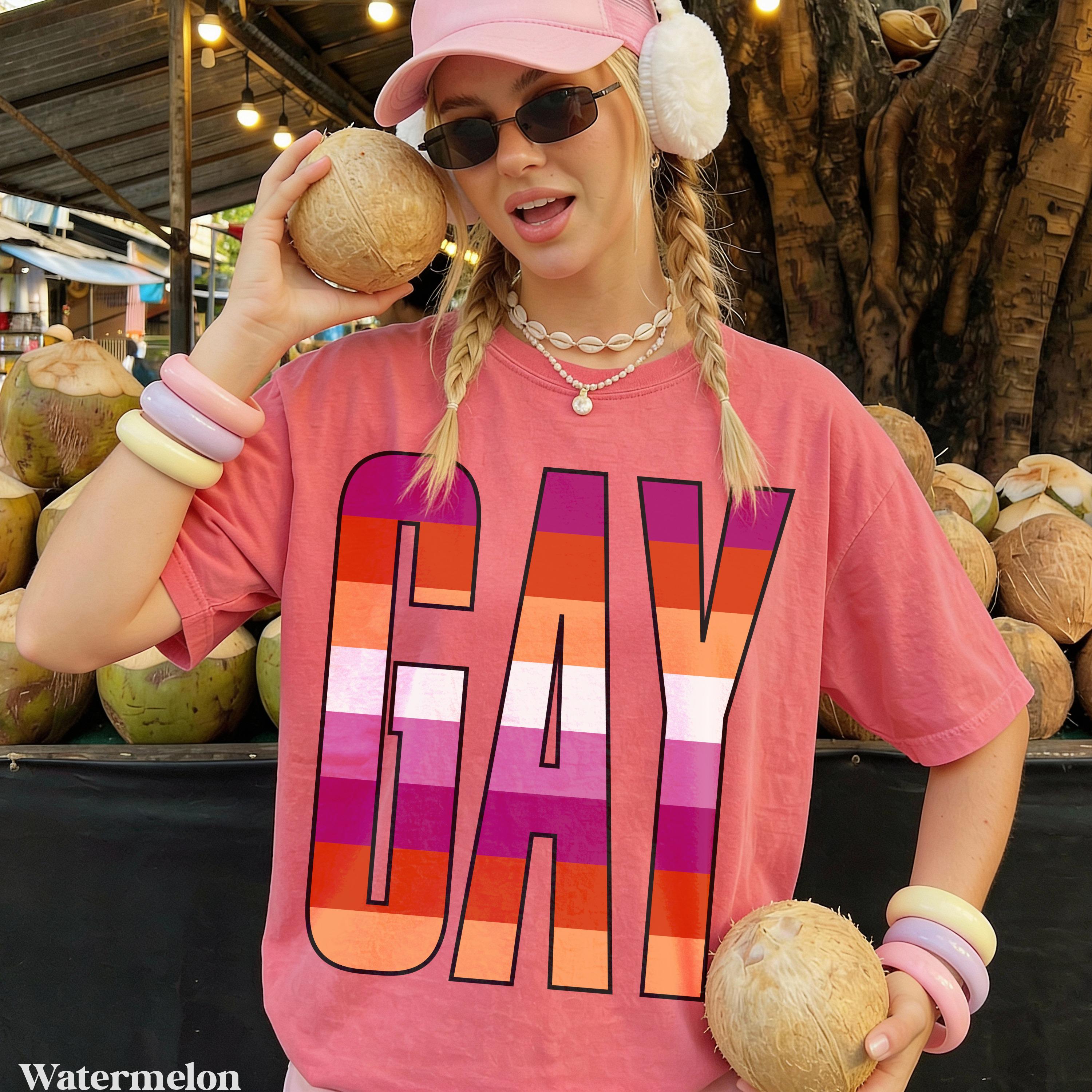 Woman in pink t-shirt with "GAY" in lesbian pride flag colors, holding coconuts.