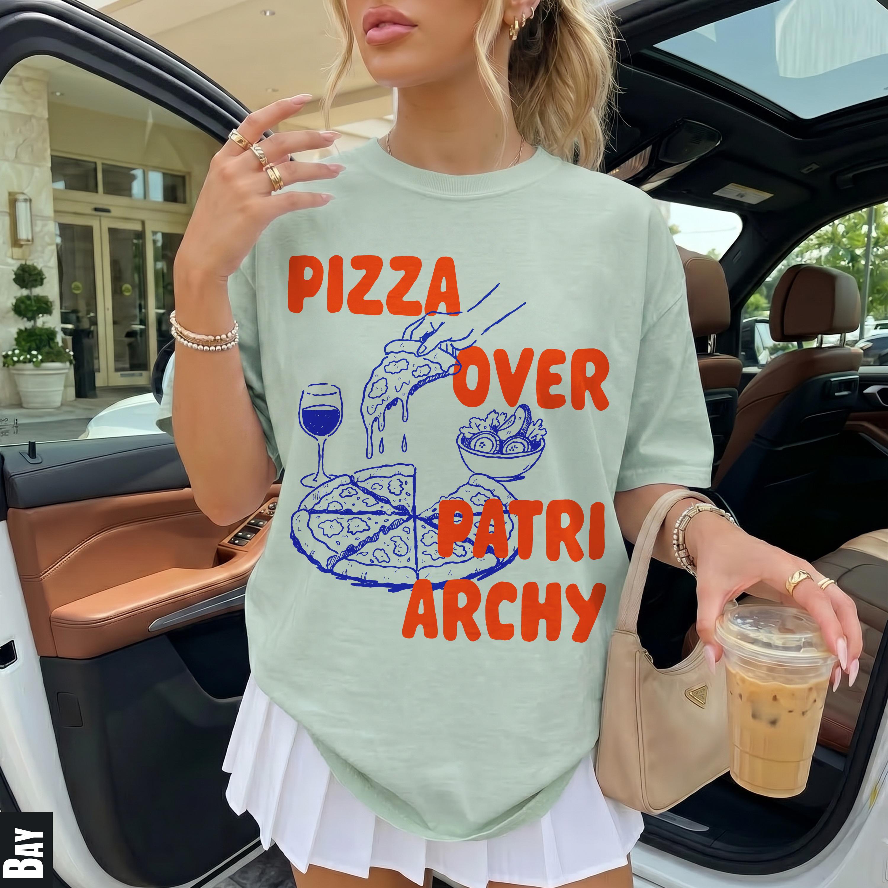 T-shirt with "Pizza Over Patriarchy" text and pizza graphic, held by a person in a car.