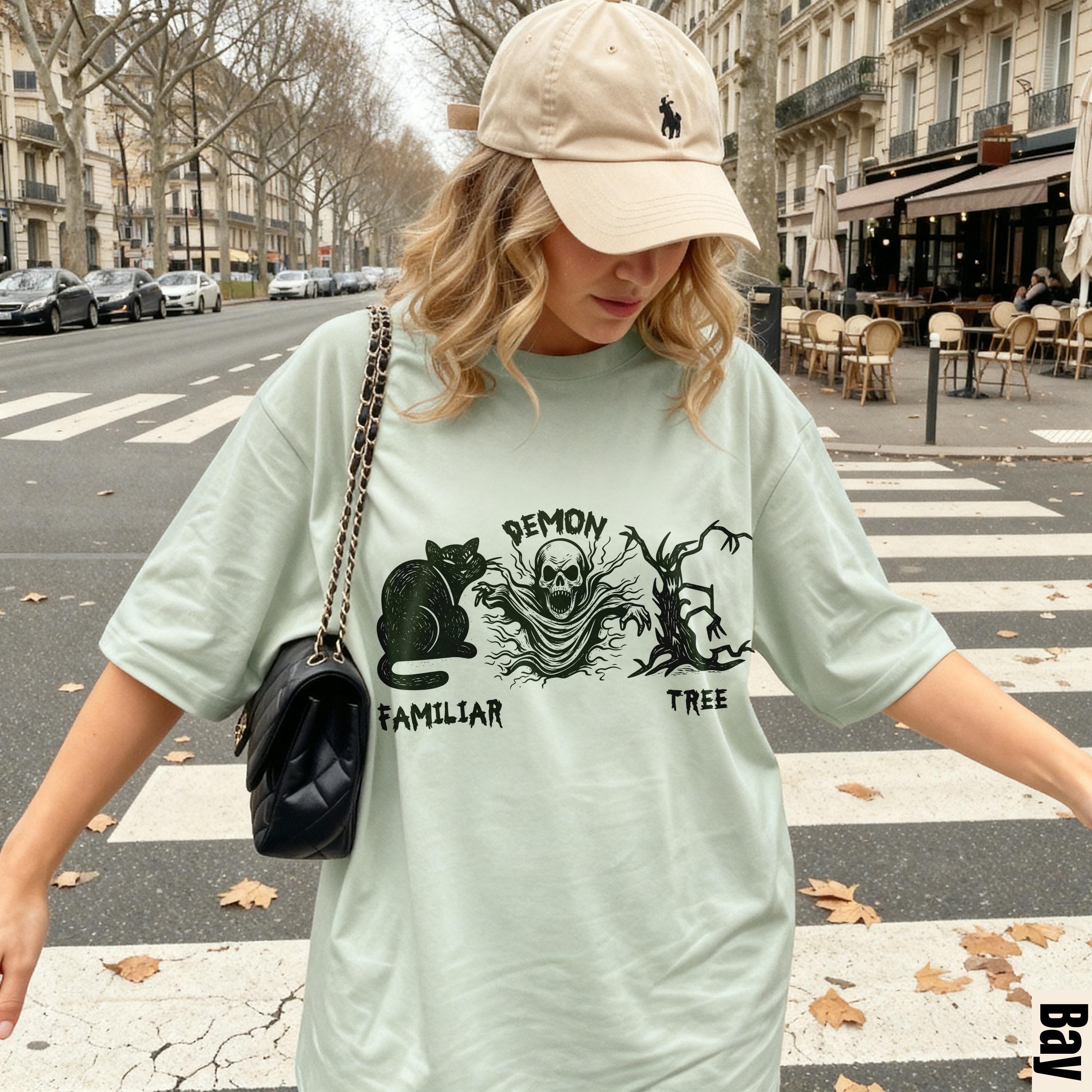 Light green t-shirt with "Demon Familiar Tree" graphic, woman wearing beige cap and black purse.