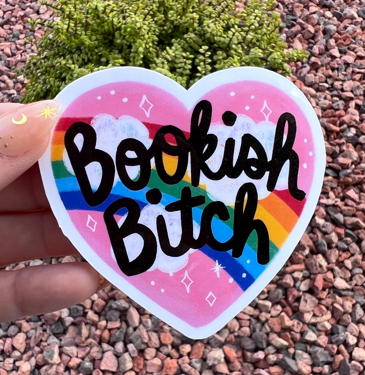 May include: A heart-shaped sticker with the words "Bookish Bitch" in black script. The sticker features a rainbow, clouds, and stars against a pink background. The sticker is held up by a hand.
