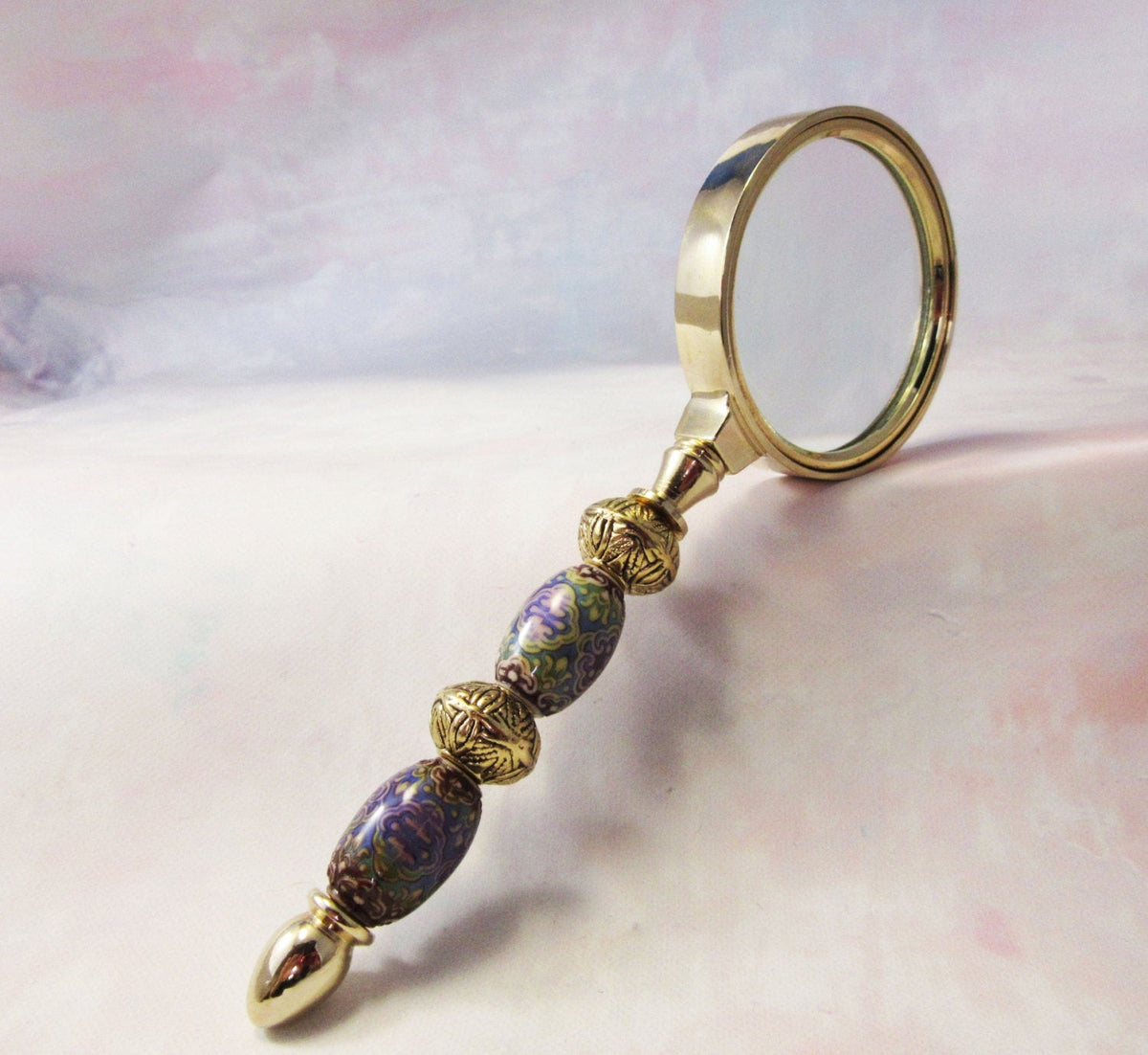 The Color Changing Magnifying Glass: Boho Office Decor features an ornate gold and purple handle with intricate patterns—ideal for mindful study and adding boho flair to your workspace. Shown resting on a soft, pastel-colored surface.