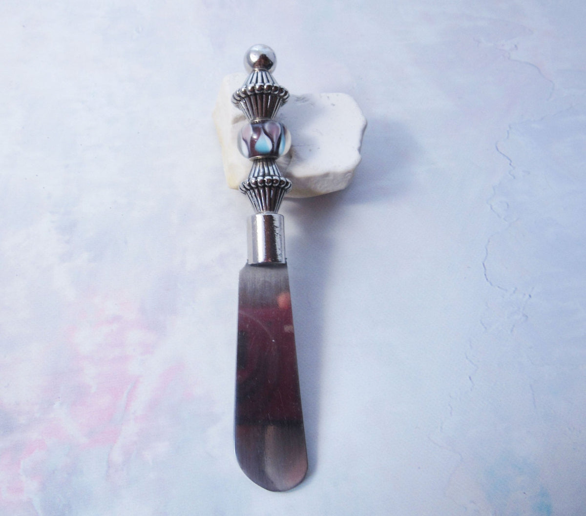 The Handcrafted Dip Spreader Knife – Glass Beads, Modern Entertaining features a silver-detailed glass bead handle and rests on white stone against a pastel background—an ideal accent for stylish gatherings.
