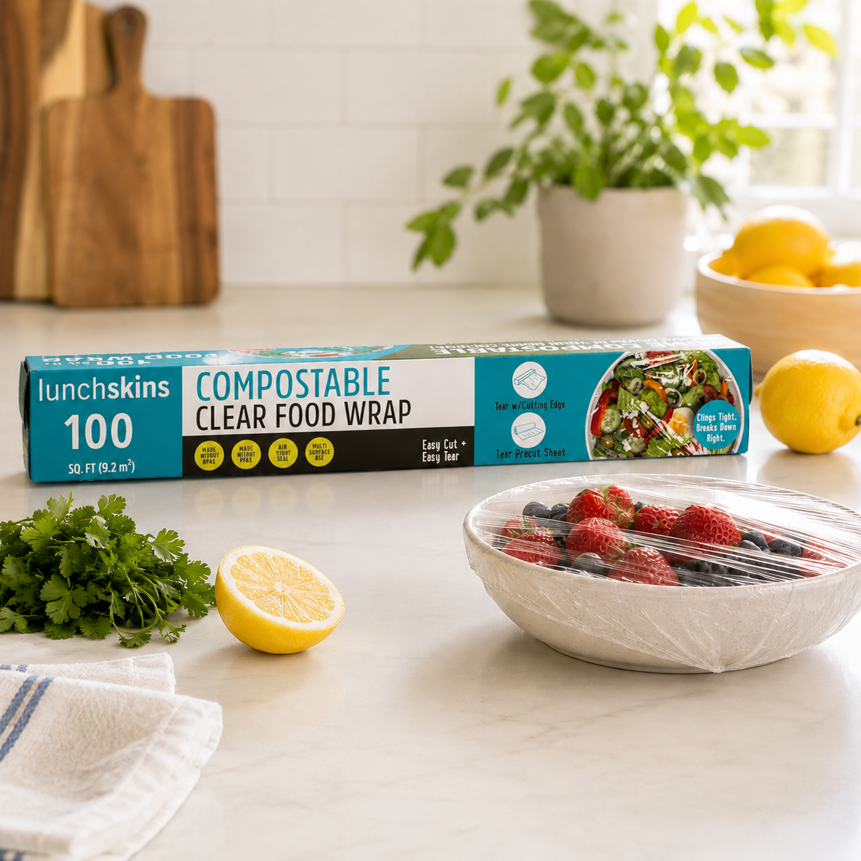 Lunchskins compostable clear food wrap box displayed on a bright kitchen counter beside a bowl of fresh berries covered with transparent compostable food wrap, lemons, and fresh herbs.