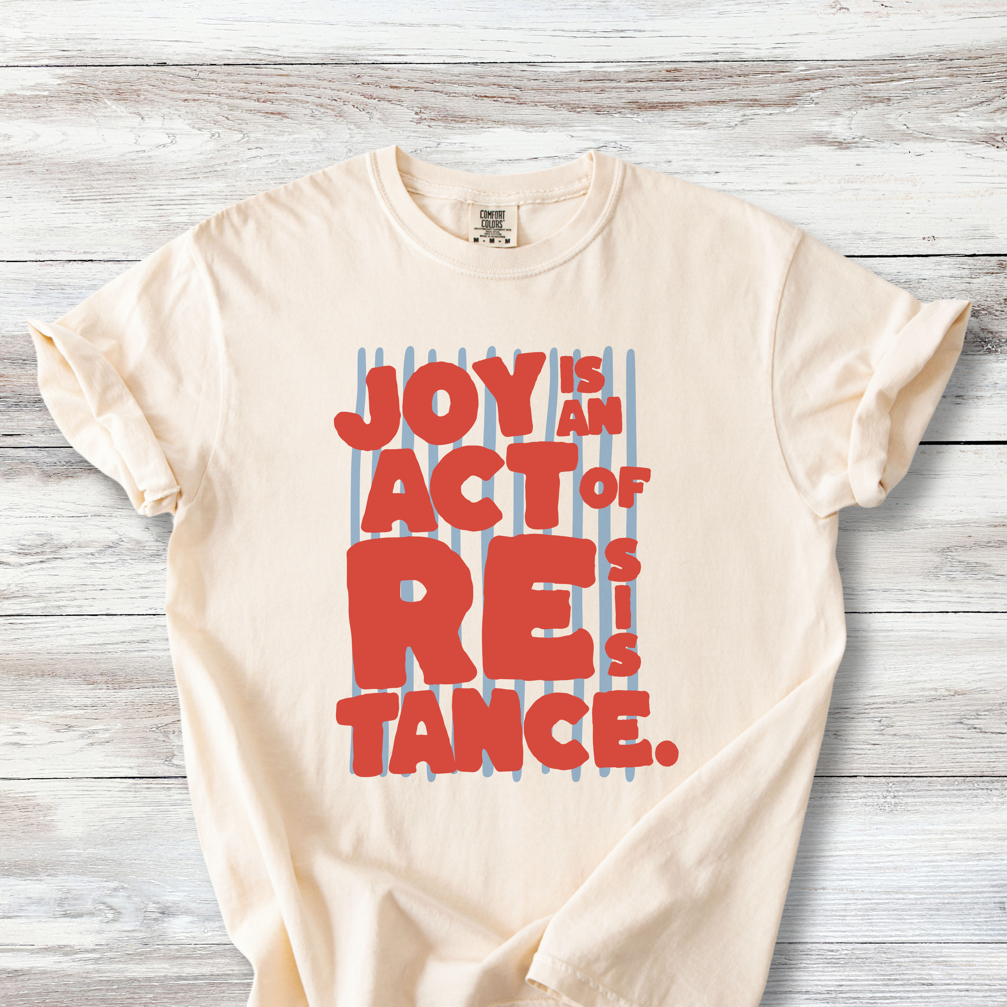 Ivory Comfort Colors t-shirt with bold red-orange chunky lettering reading "Joy Is an Act of Resistance" layered over a muted slate blue vertical stripes