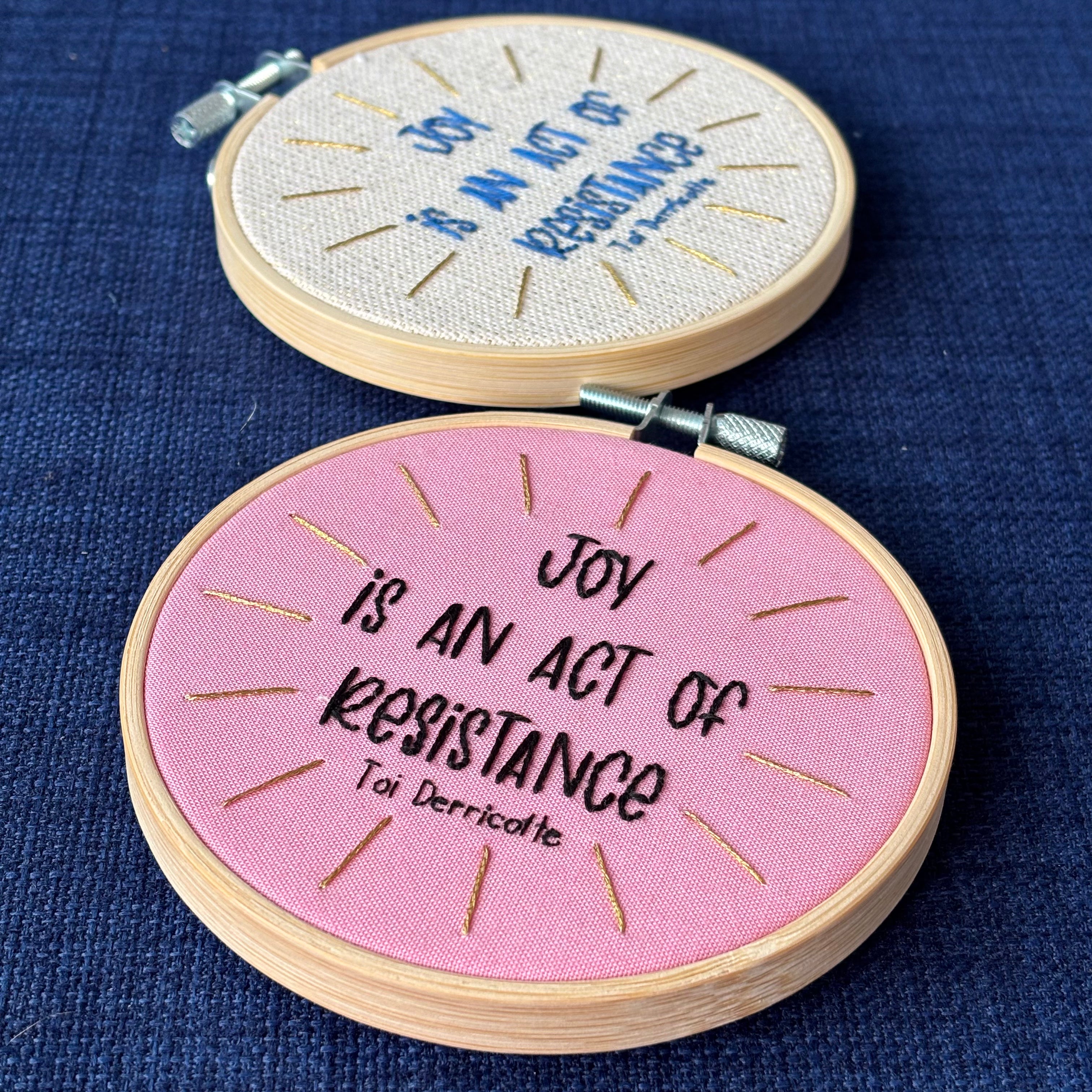 Two hand embroidery hoops that read, "Joy is an act of resistance" - Toi Derricotte. Text is a handwriting font and hand embroidered in black embroidery thread. Words are centered in a burst of metallic gold rays. Fabric is light pink cotton. Framed in light wood embroidery hoop. In the background is a second hoop with the same message, with the words in blue thread, gold metallic rays, and a sparking cream linen fabric.