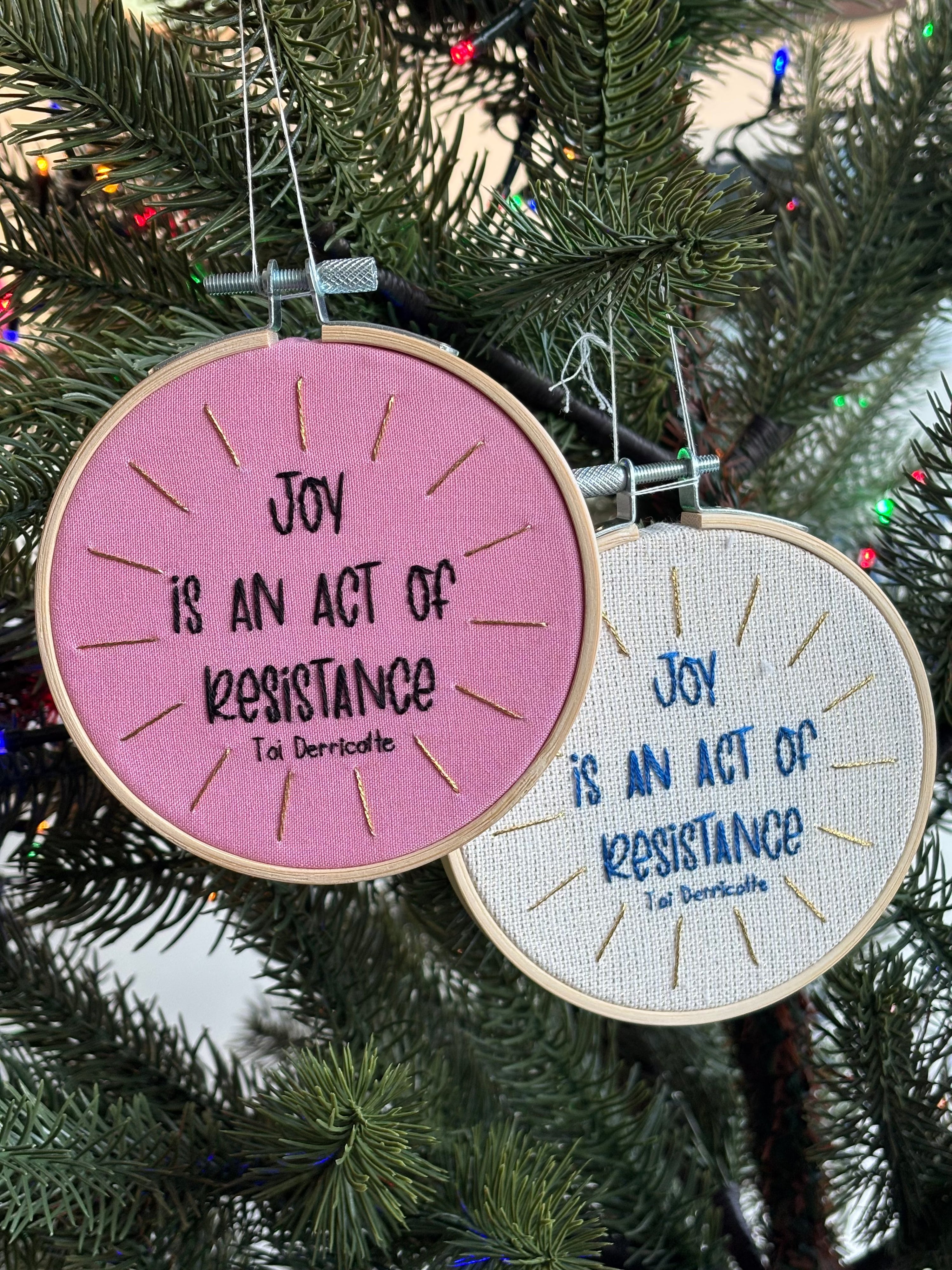 Two hand embroidery hoops that read, "Joy is an act of resistance" - Toi Derricotte. Hoops are hanging on a Christmas tree as Christmas ornaments. Text is a handwriting font. One ornament is hand embroidered in black embroidery thread. Words are centered in a burst of metallic gold rays. Fabric is light pink cotton. Framed in light wood embroidery hoop. In a second hoop with the same message, the words are sewn in blue thread with gold metallic rays on a sparking cream linen fabric.