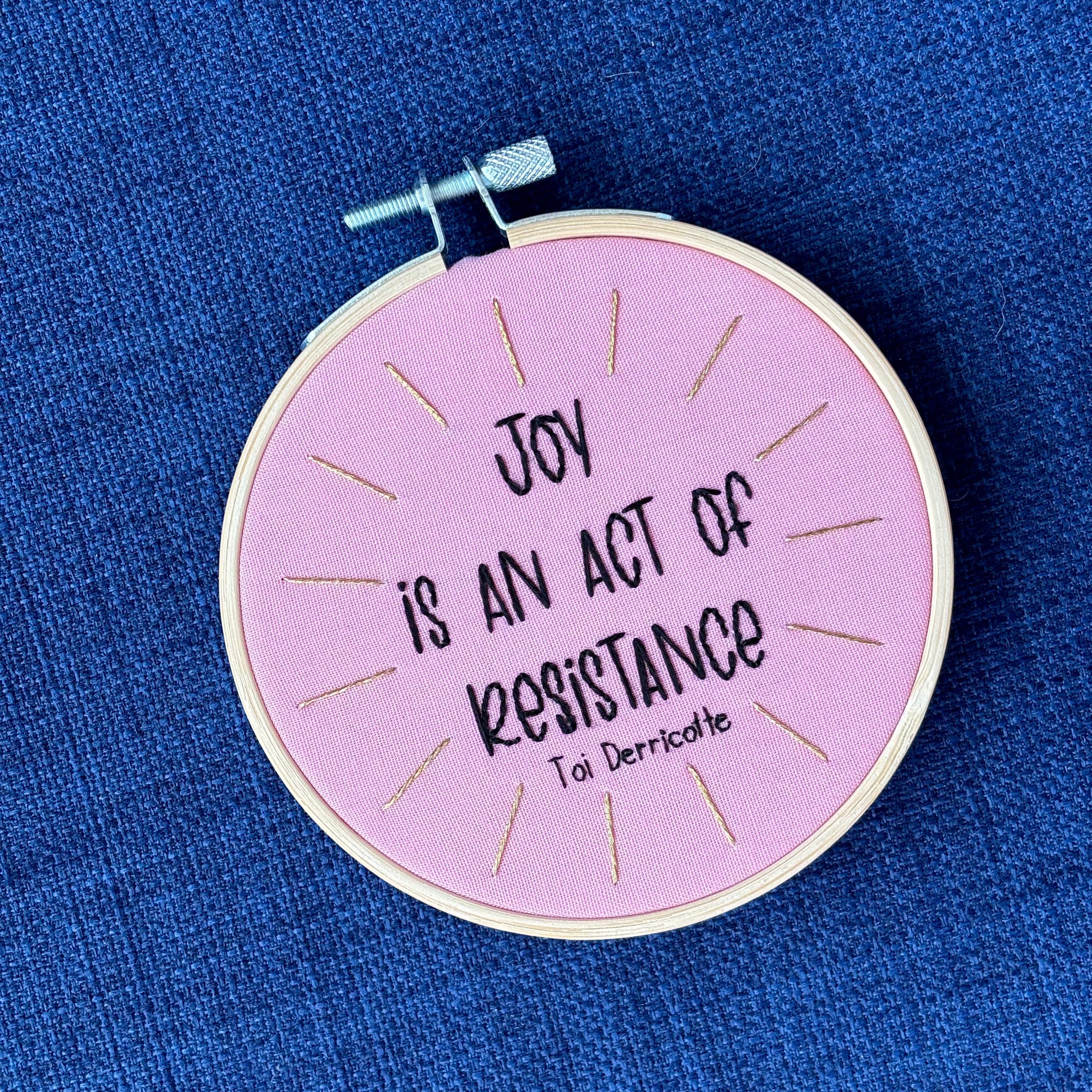 "Joy is an act of resistance" - Toi Derricotte. Text is a handwriting font and hand embroidered in black embroidery thread. Words are centered in a burst of golden, metallic rays. Fabric is light pink cotton. Framed as wall art or christmas ornament in light wood embroidery hoop.