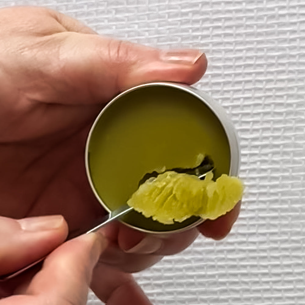 A close-up of Nordic Meadow Balm. Hand holding a small round metal tin filled with a green balm or wax, while a spoon lifts a textured, crumbly olive-green piece from the tin against a white textured background.