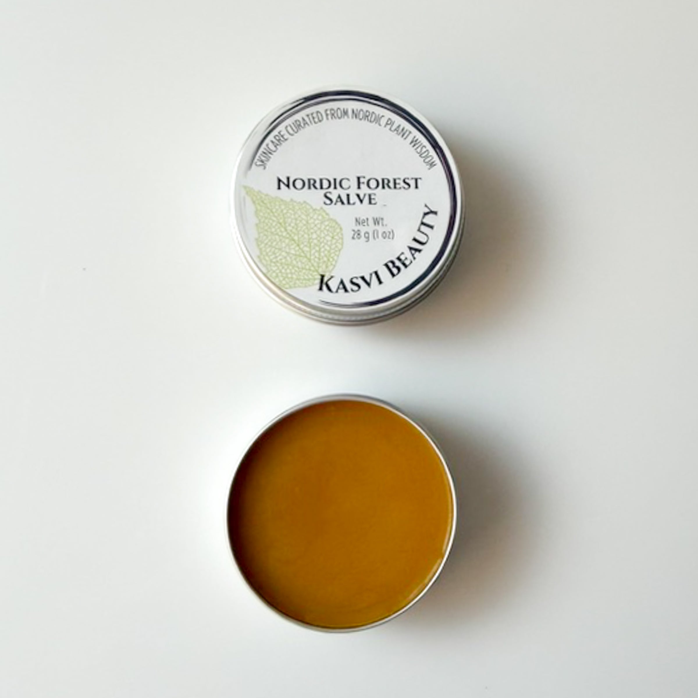 An open round metal tin of Kasvi Beauty Nordic Forest Salve, showing the golden-brown balm inside, with the lid placed above on a clean white background.