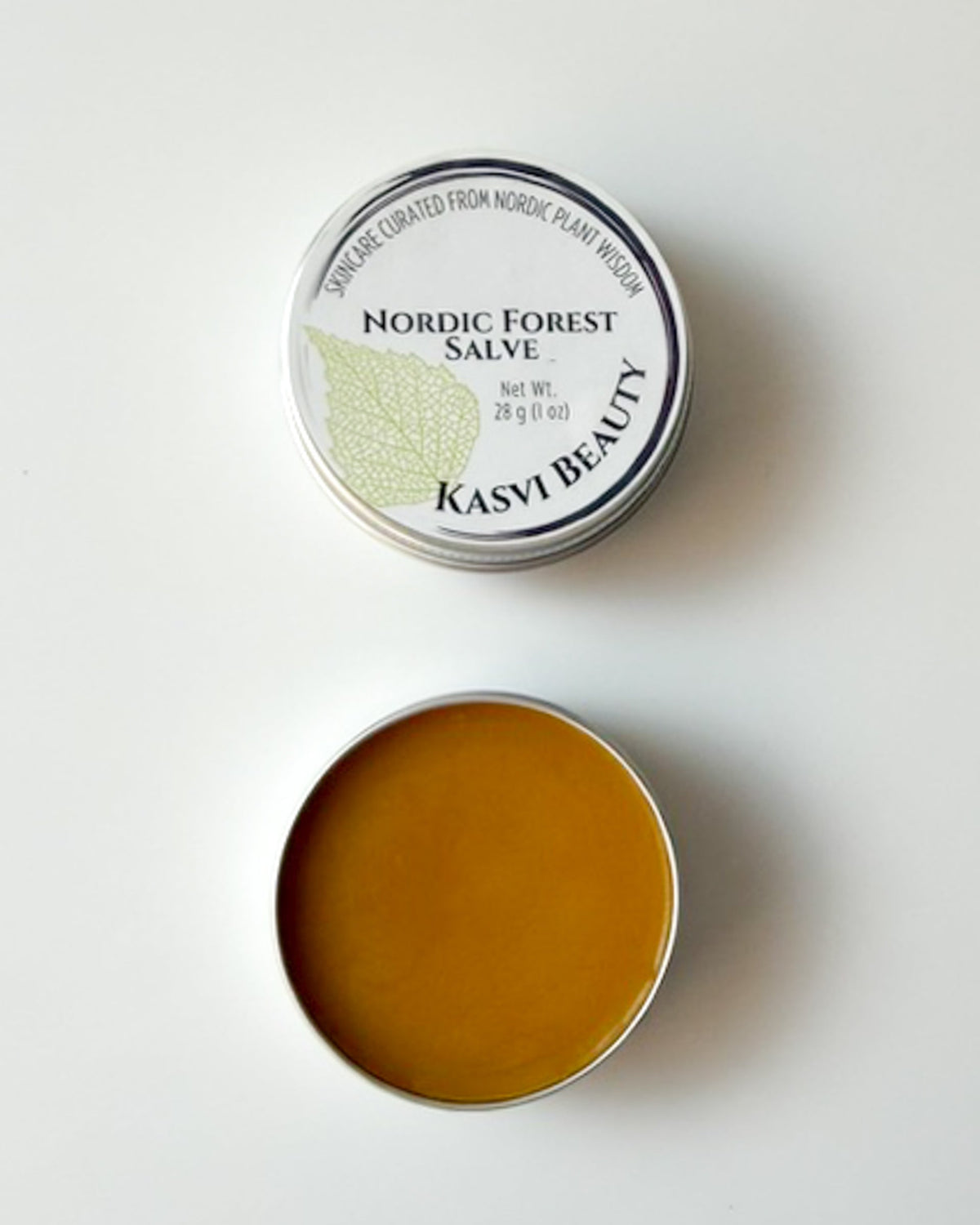 An open round metal tin of Kasvi Beauty Nordic Forest Salve, showing the golden-brown balm inside, with the lid placed above on a clean white background.