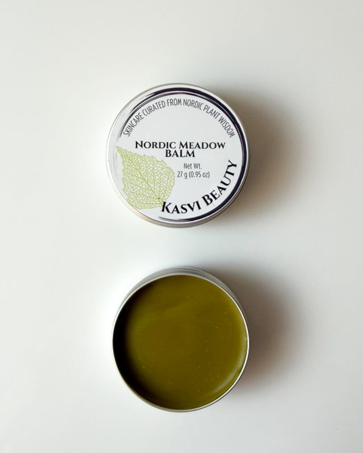 An open round metal tin of Kasvi Beauty Nordic Meadow Balm, showing the olive-green balm inside, positioned below its closed lid which features a white label with a green birch leaf graphic.
