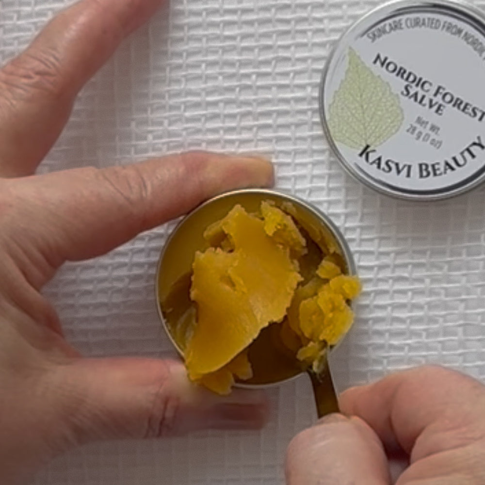 A close-up of hands holding an open metal tin of Kasvi Beauty Nordic Forest Salve, showing the yellow balm inside, while a spoon lifts a textured, crumbly golden yellow piece from the tin with the branded lid resting nearby on a textured white surface.