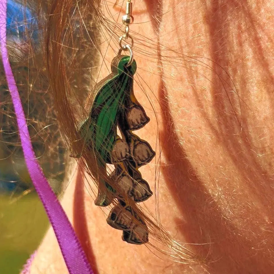 Close-up of hand-painted wooden lily of the valley dangle earrings with green leaves and white bell flowers and hypoallergenic sterling silver hooks, delicate floral statement jewelry worn on ear