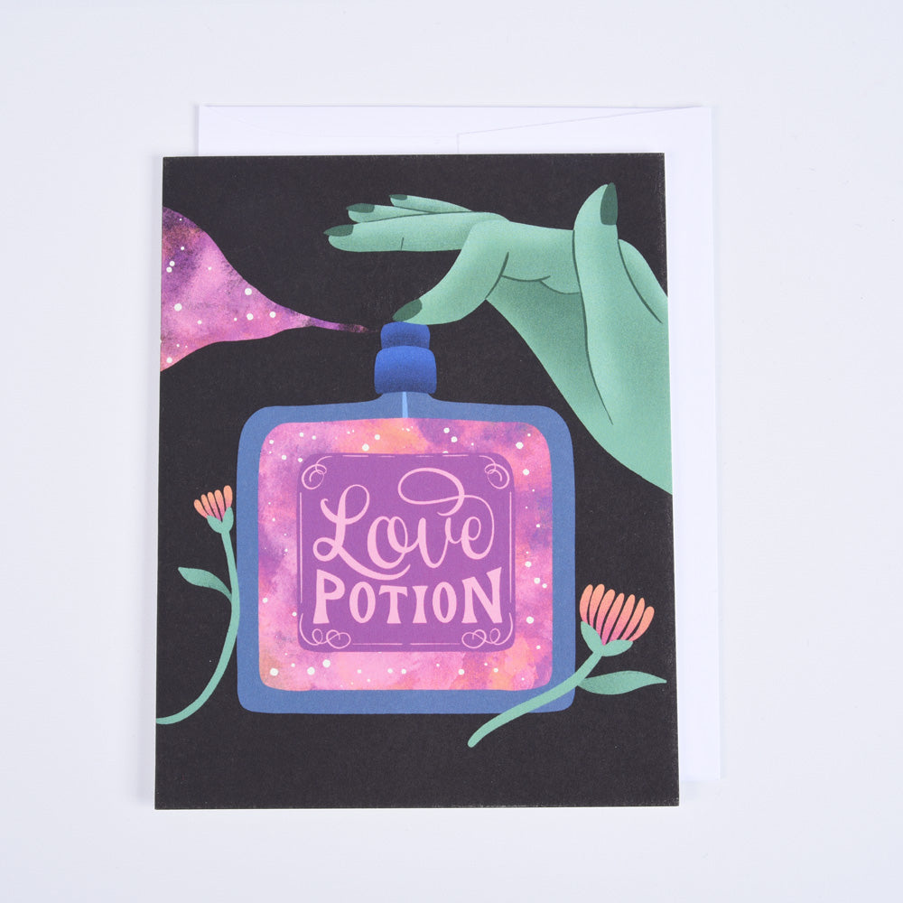 A folded greeting card with a colorful illustration of a love potion bottle, stars, and a green hand, with the phrase &