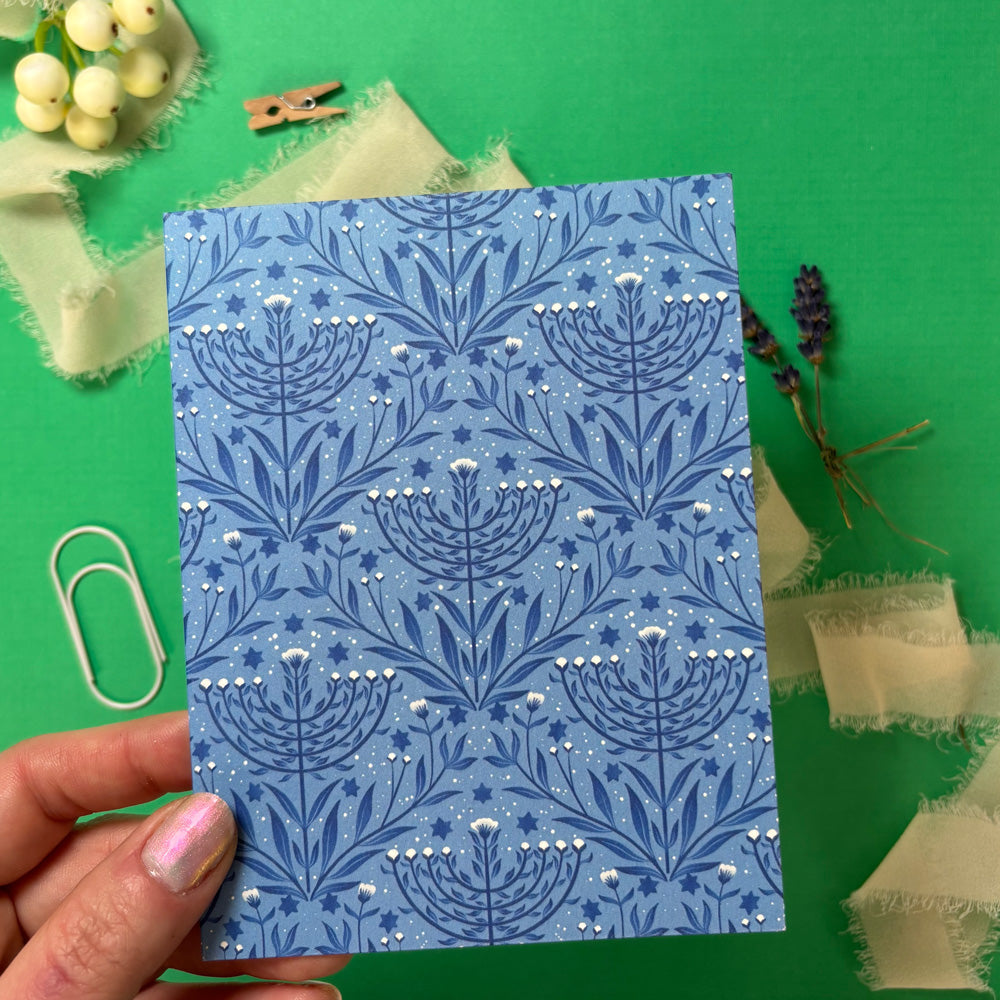 Blue patterned menorah card held by a hand on a green background with decorative items.