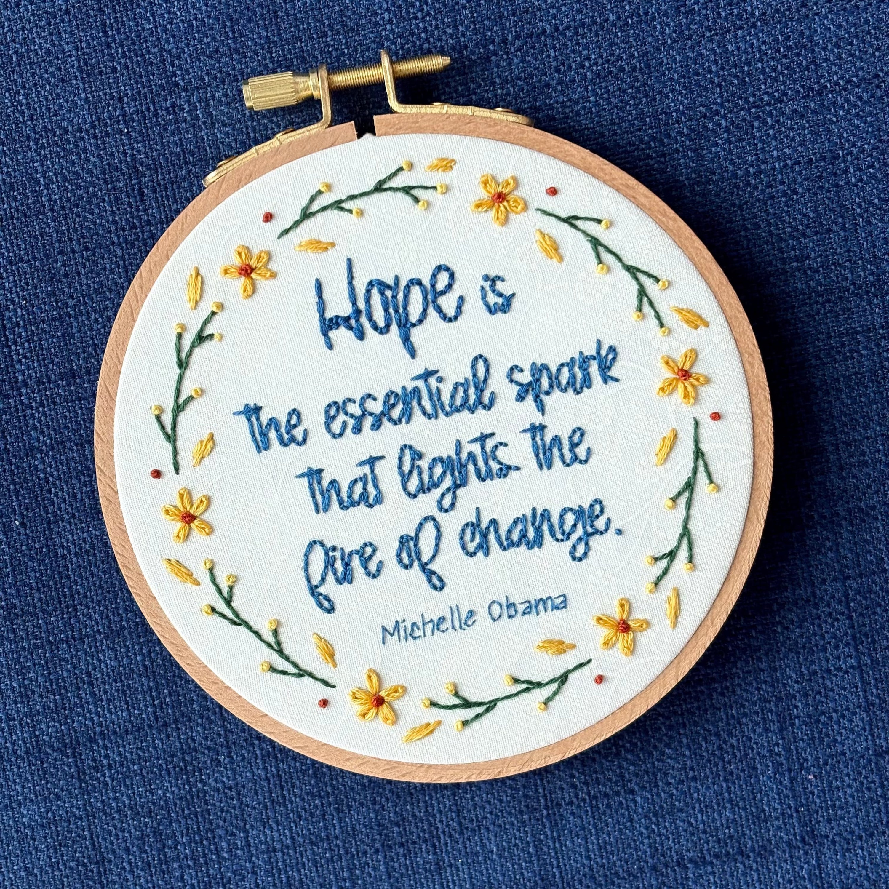 "Hope is the essential spark that lights the fire of change. Michelle Obama" Quote hand embroidered in dark blue. Text is surrounded by floral wreath of golden yellow sunflowers and dark green stems with light yellow buds. Fabric is white-on-white cotton. Displayed as wall art in a light wooden embroidery hoop.