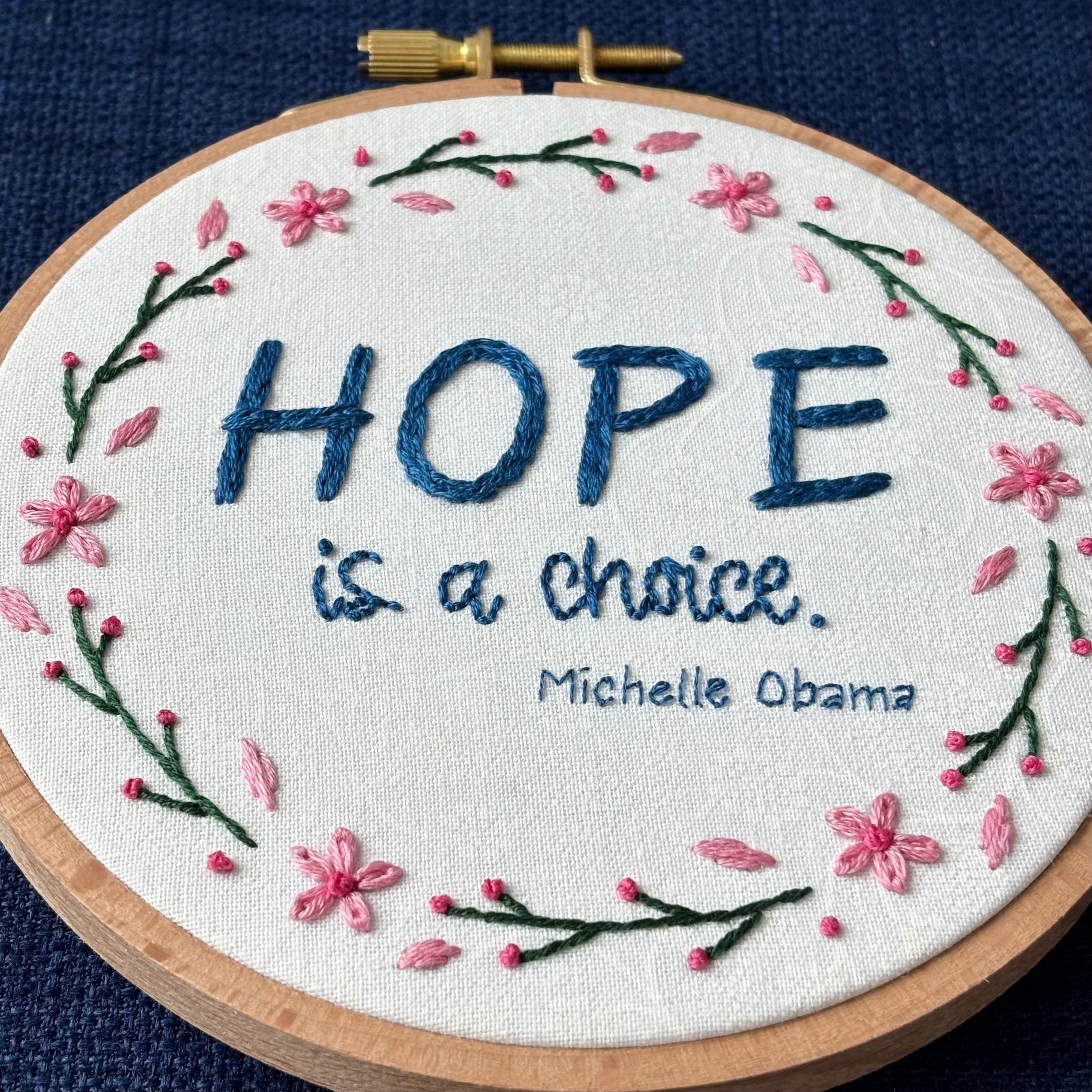 "Hope is a choice. Michelle Obama" Quote hand embroidered in dark blue. Text is surrounded by floral wreath of pink cherry blossom flowers and stems with buds. Fabric is white-on-white cotton. Displayed as wall art in a light wooden embroidery hoop.