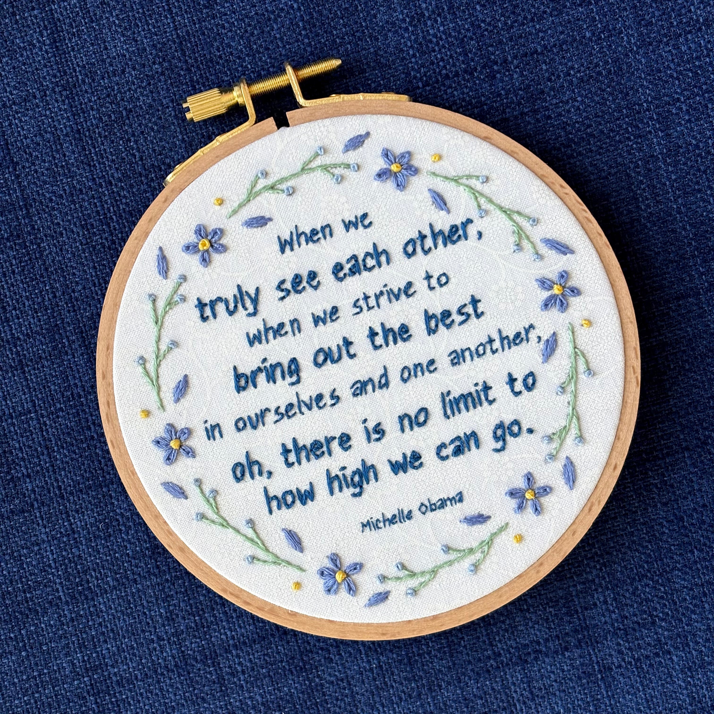 "When we truly see each other, when we strive to bring out the best in ourselves and one another, oh, there is no limit to how high we can go. Michelle Obama." Quote hand embroidered in dark blue. Text is surrounded by floral wreath of periwinkle blue forget-me-not flowers and light green stems with light blue buds. Fabric is white-on-white cotton. Displayed as wall art in a light wooden embroidery hoop.