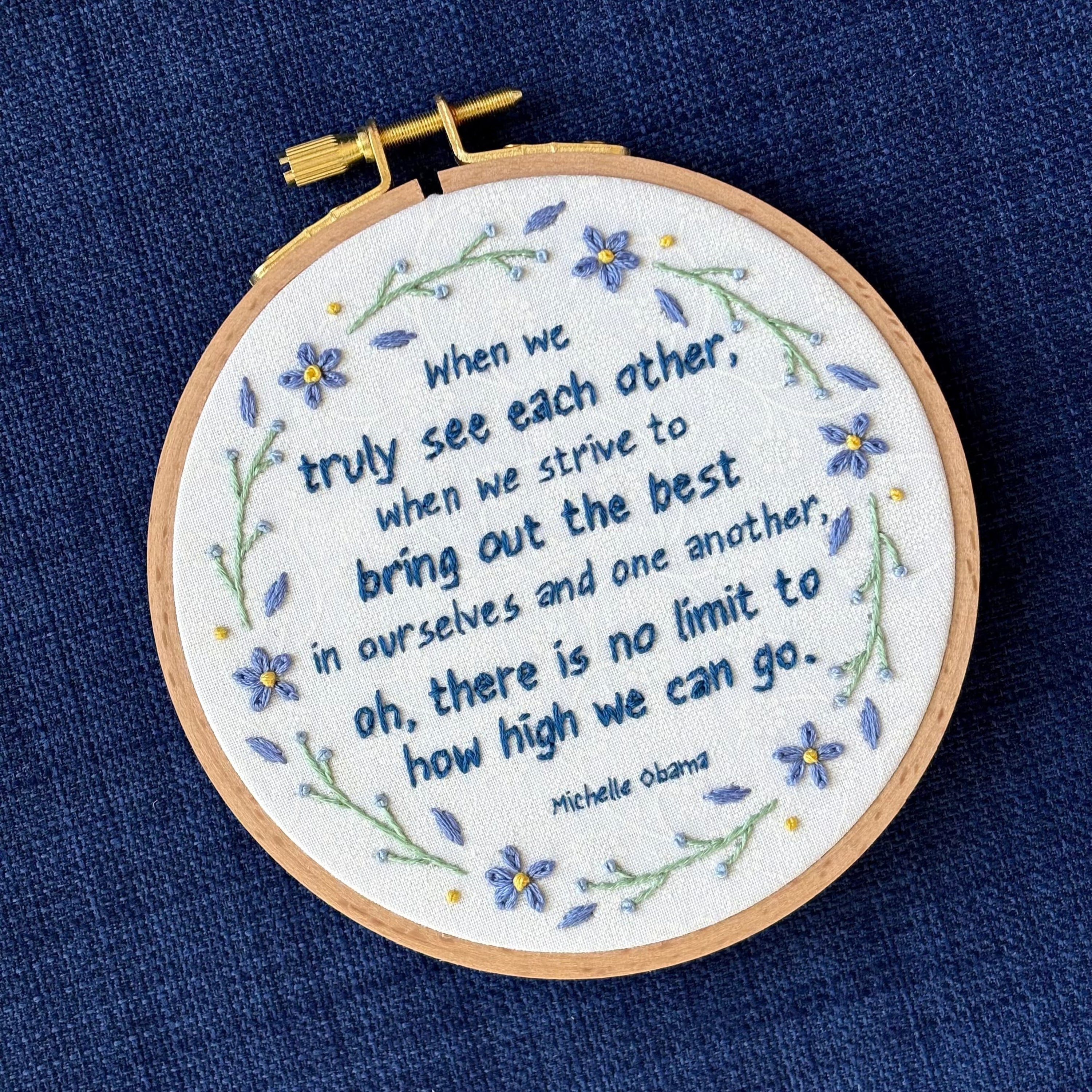 "When we truly see each other, when we strive to bring out the best in ourselves and one another, oh, there is no limit to how high we can go. Michelle Obama." Quote hand embroidered in dark blue. Text is surrounded by floral wreath of periwinkle blue forget-me-not flowers and light green stems with light blue buds. Fabric is white-on-white cotton. Displayed as wall art in a light wooden embroidery hoop.