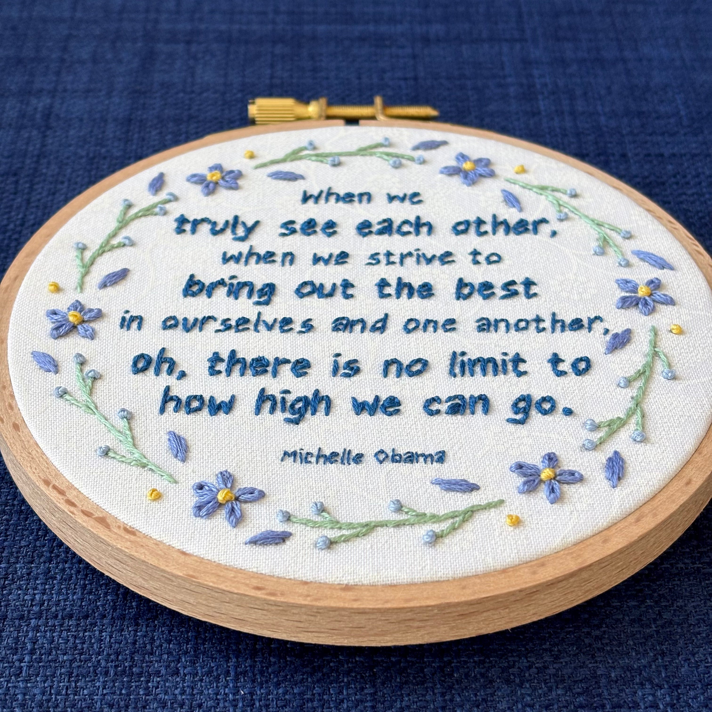 "When we truly see each other, when we strive to bring out the best in ourselves and one another, oh, there is no limit to how high we can go. Michelle Obama." Quote hand embroidered in dark blue. Text is surrounded by floral wreath of periwinkle blue forget-me-not flowers and light green stems with light blue buds. Fabric is white-on-white cotton. Displayed as wall art in a light wooden embroidery hoop.