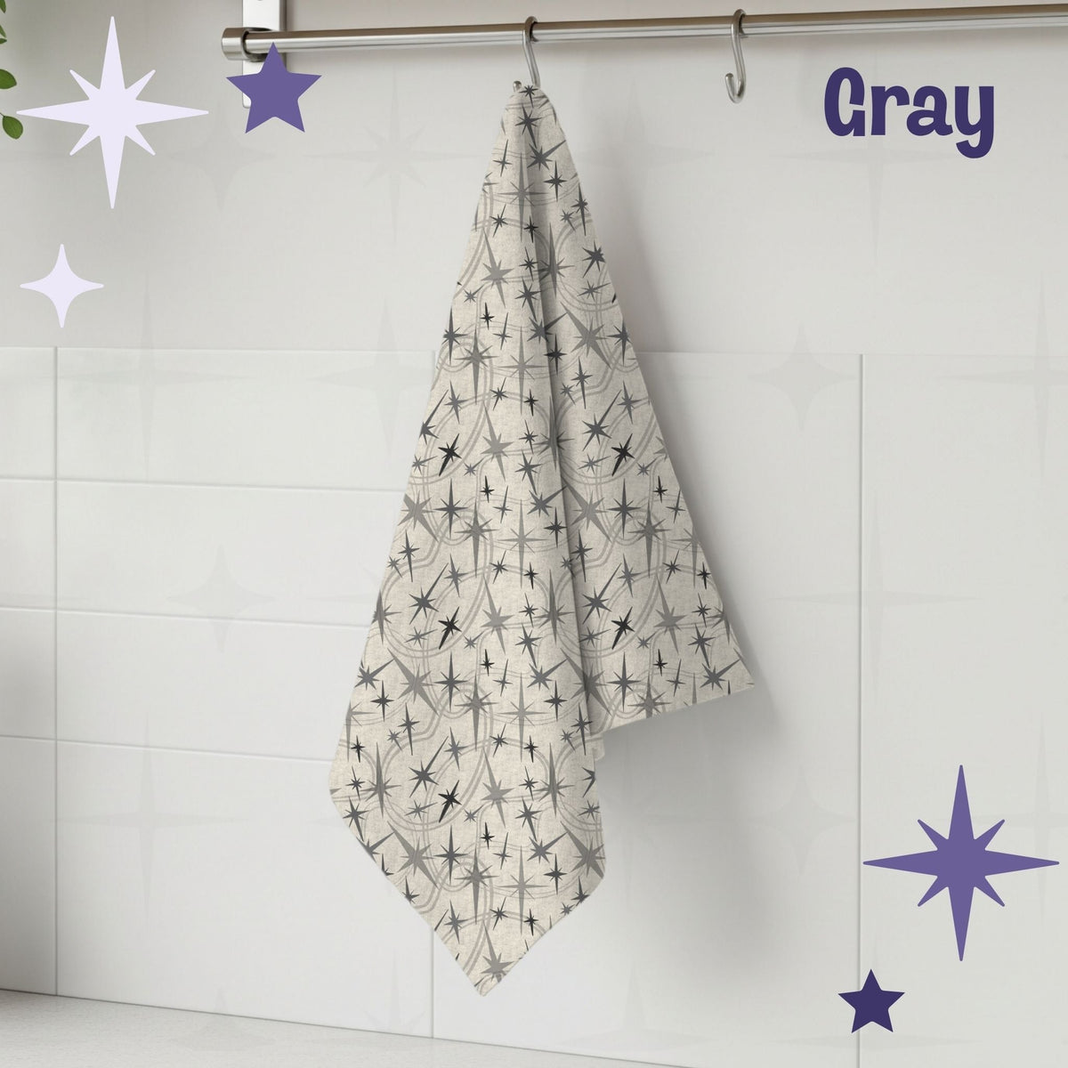 Warm gray atomic starburst mid century modern tea towel hanging from a chrome hook against white subway tile, showing full pattern of charcoal and gray starburst motifs on a natural oatmeal cotton twill background