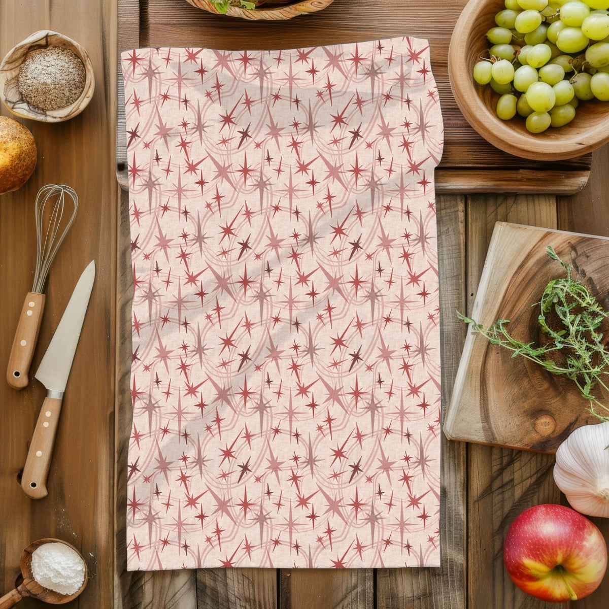 Overhead flat lay of a mid century modern tea towel in the dusty rose colorway laid on a rustic wood surface surrounded by kitchen props including a whisk, grapes, apple, garlic, and fresh herbs