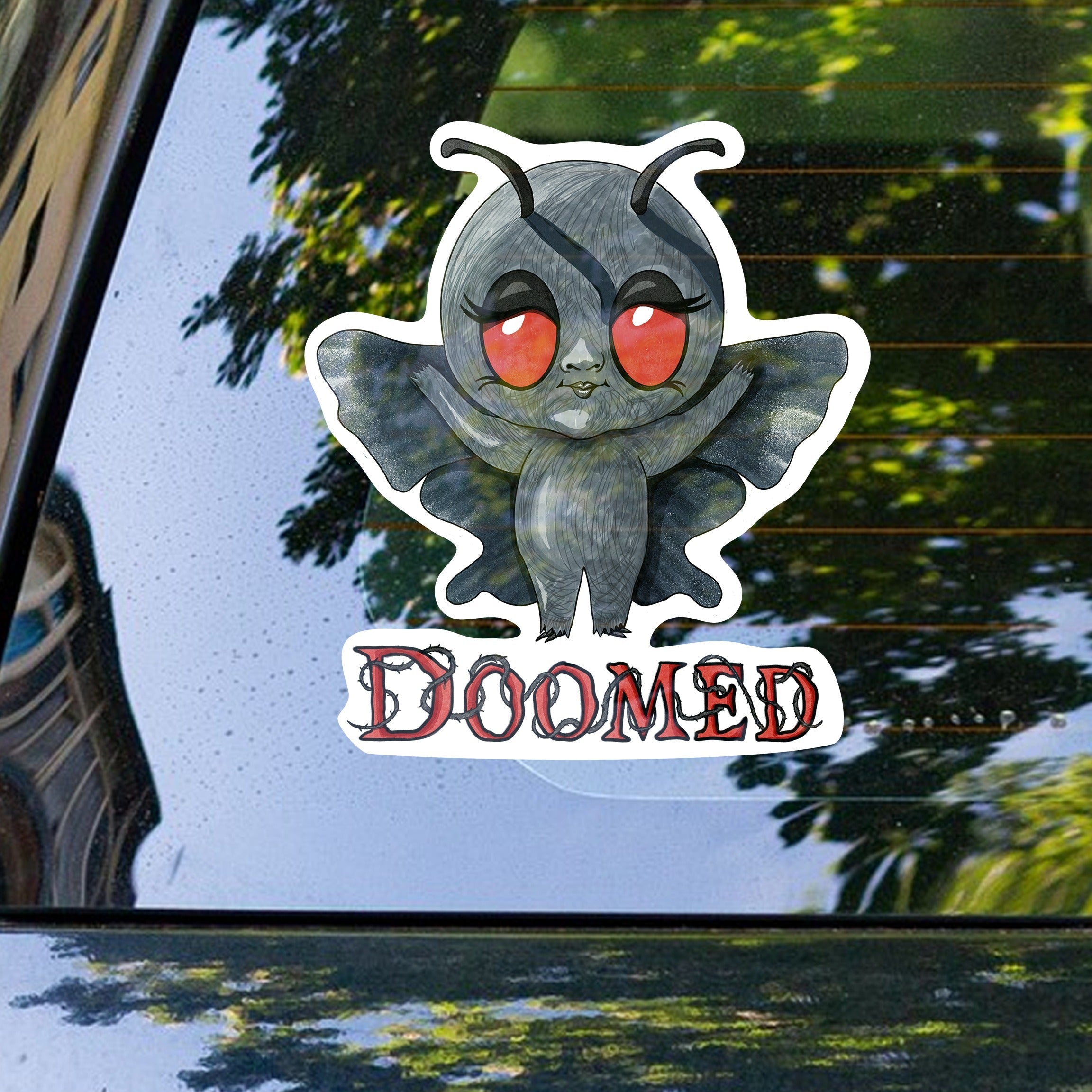 Mothman cryptid sticker on the rear window of a car with trees reflected behind