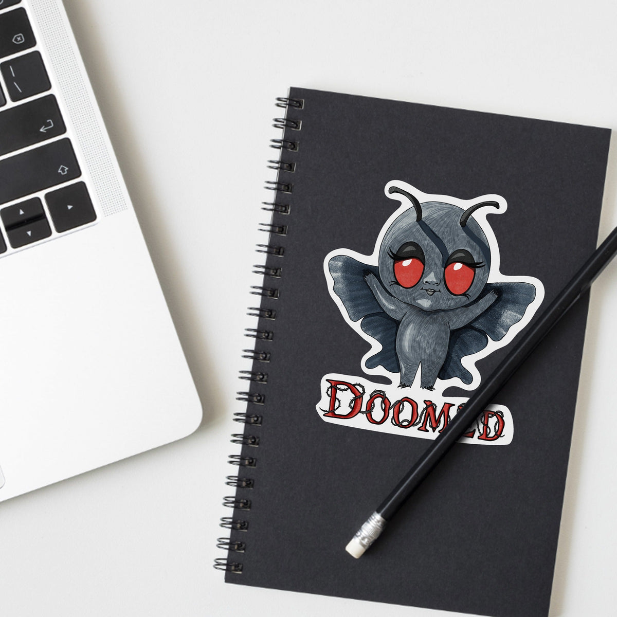 Chibi Mothman cryptid sticker on a black spiral notebook beside a laptop and pencil