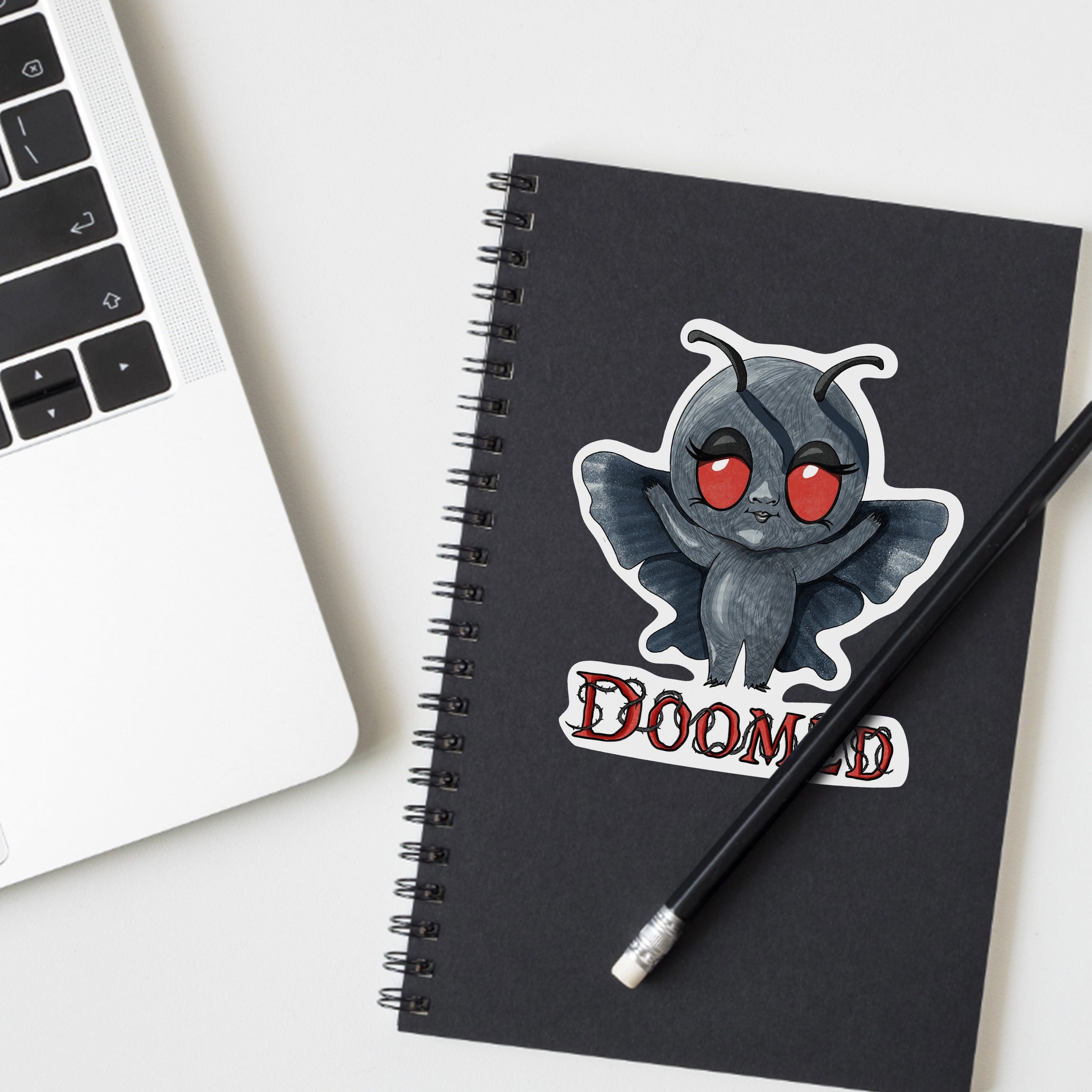 Chibi Mothman cryptid sticker on a black spiral notebook beside a laptop and pencil