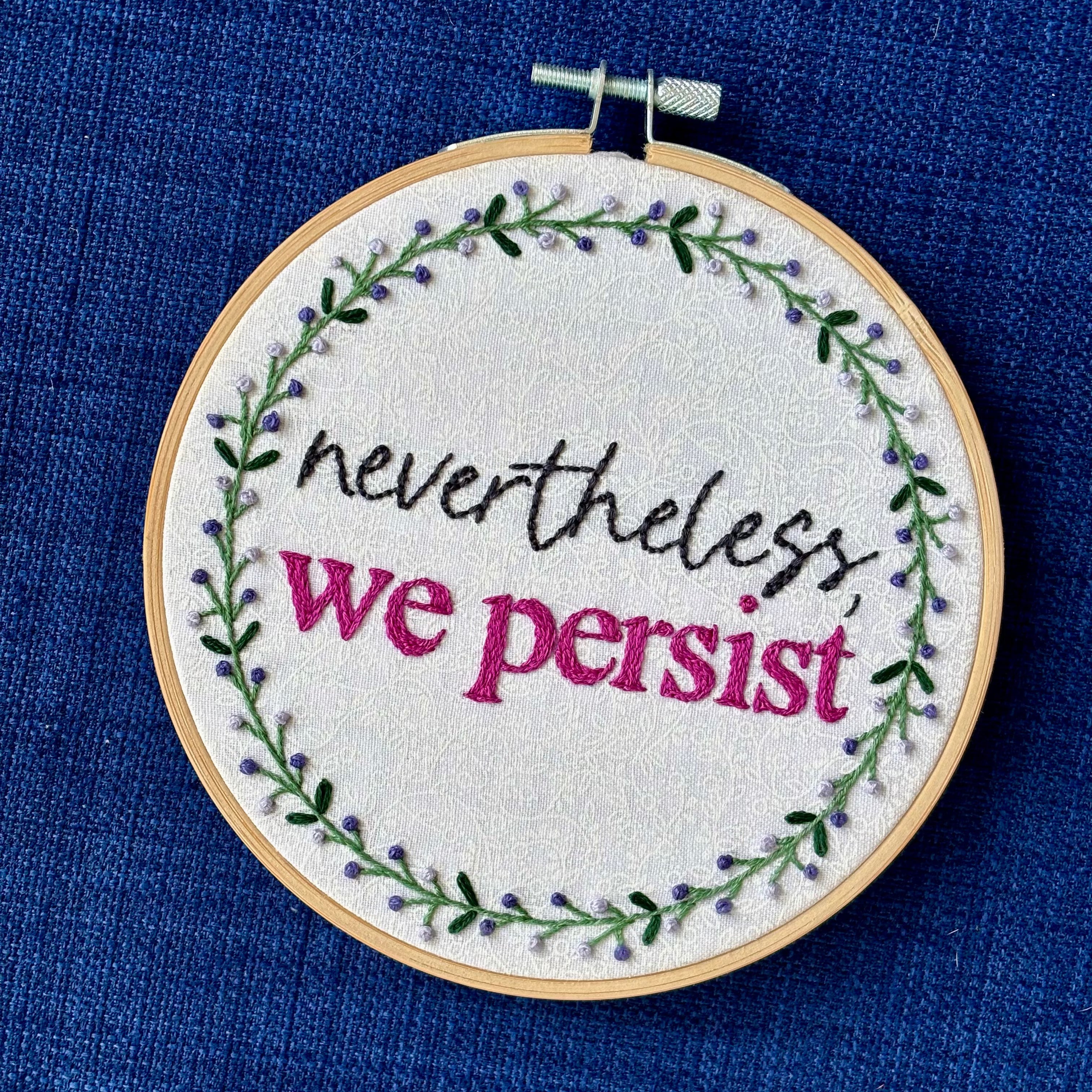 "Nevertheless, we persist." Nevertheless hand embroidered in a script font in dark gray. "we persist" is in Times New Roman and sewn bright fuchsia embroidery thread. Quote surrounded with a floral wreath with purple and lavender flower buds and green leaves. Stitched on white-on-white cotton. Wall art framed in light wood embroidery hoop.