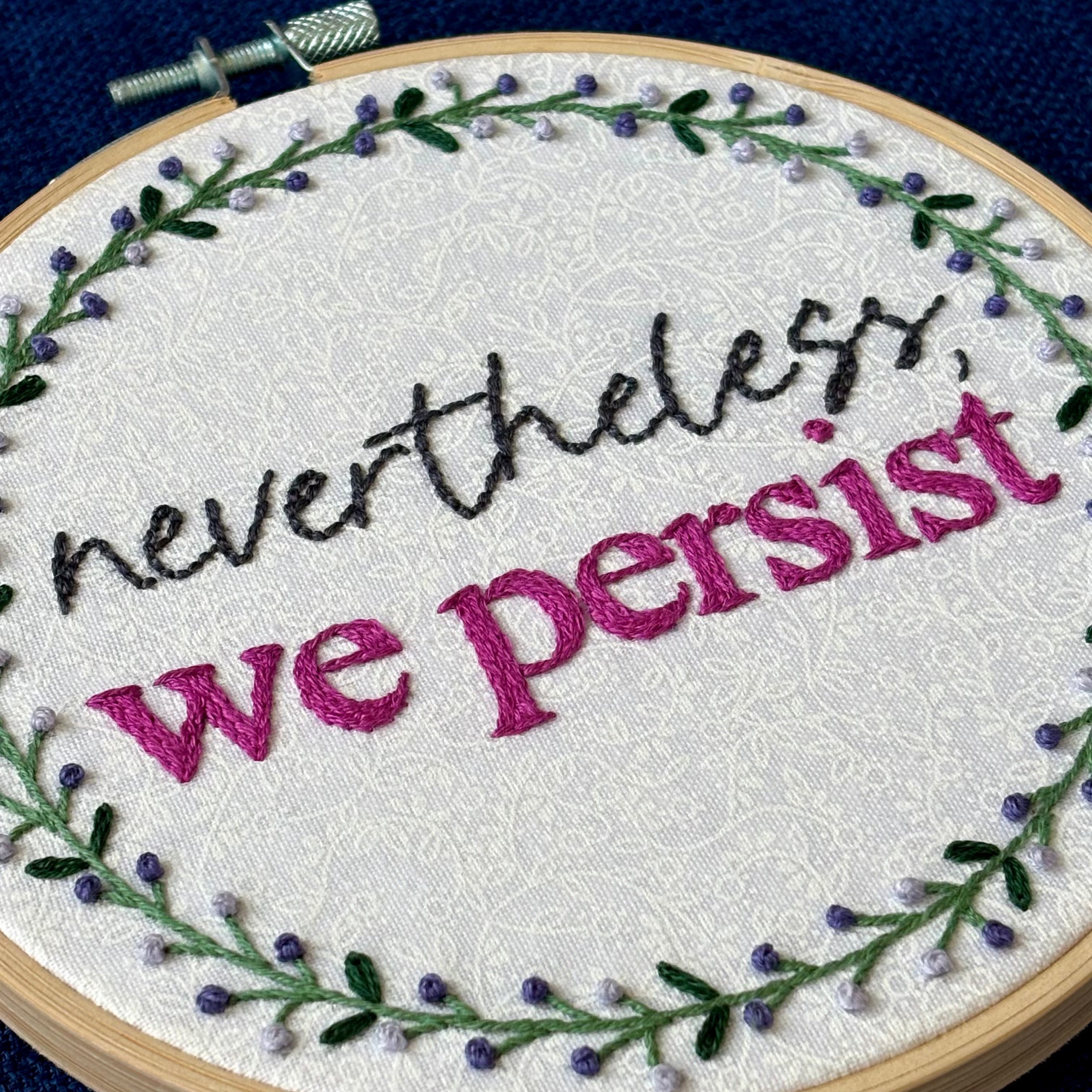 "Nevertheless, we persist." Nevertheless hand embroidered in a script font in dark gray. "we persist" is in Times New Roman and sewn bright fuchsia embroidery thread. Quote surrounded with a floral wreath with purple and lavender flower buds and green leaves. Stitched on white-on-white cotton. Wall art framed in light wood embroidery hoop.