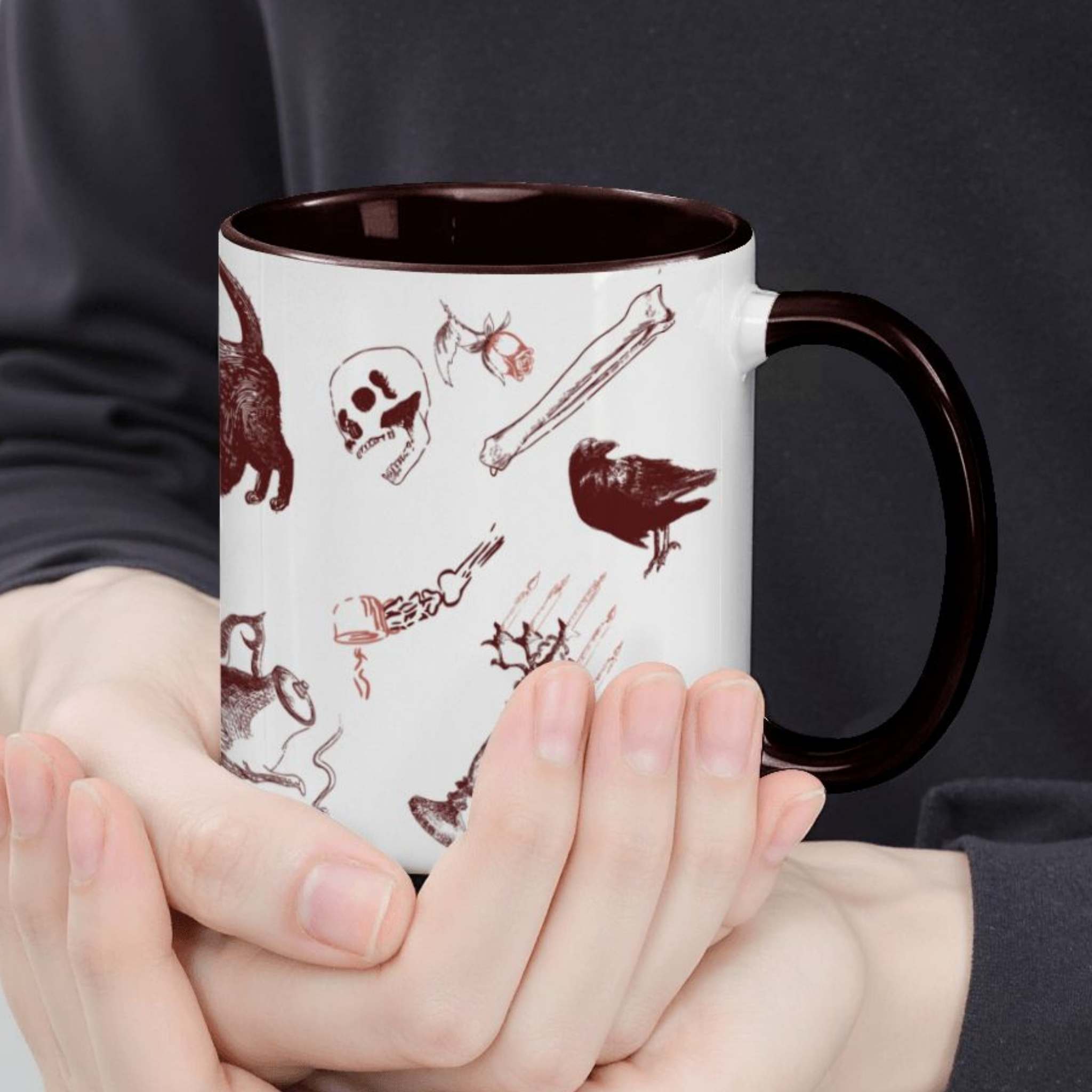Hands cradling the oxblood skull and bones mug, close view of red motifs including a skull, a raven, and a skeletal hand with a steaming teacup