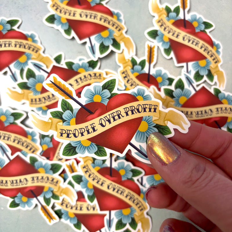 hand holding a sticker of an arrow piercing a heart that says "people over profit"