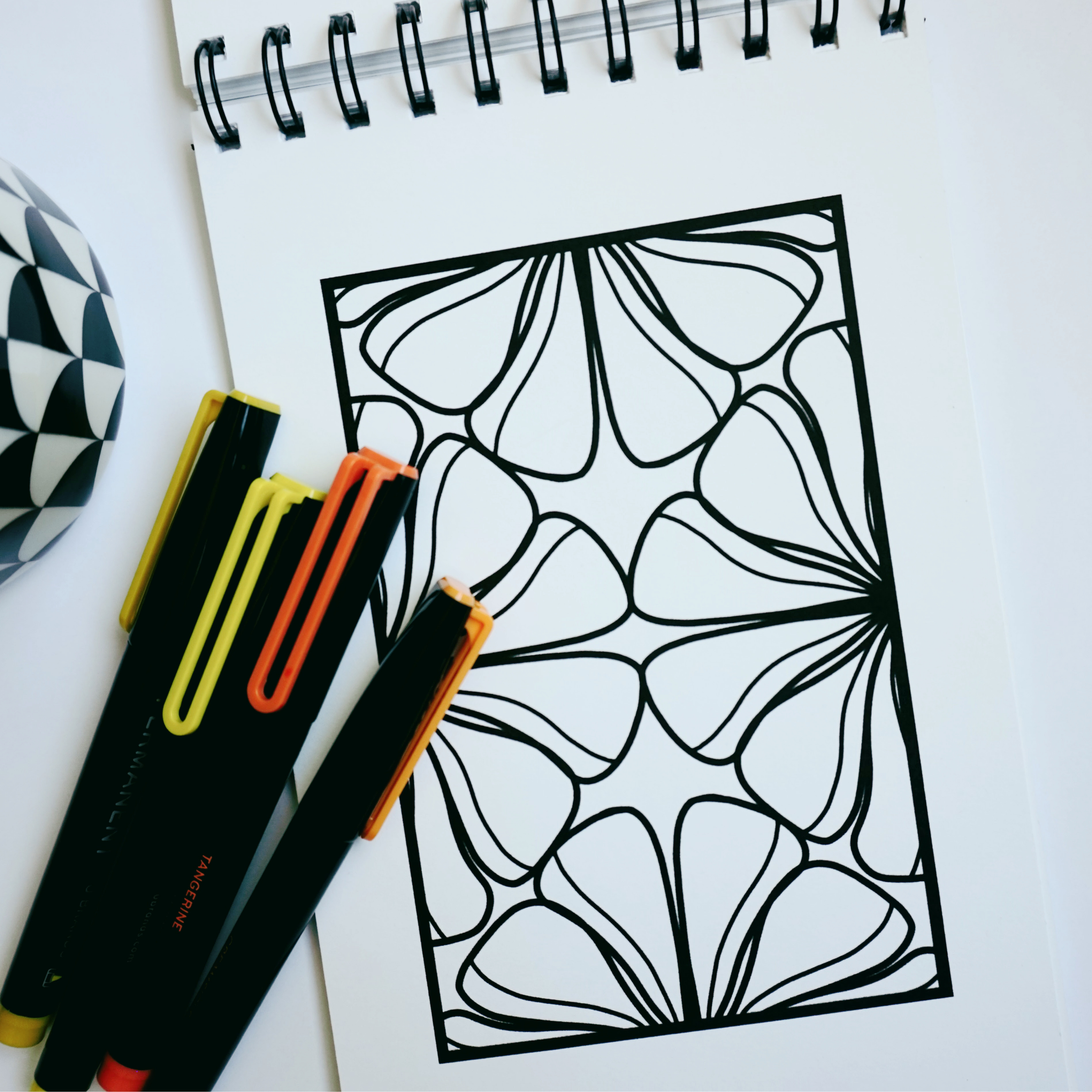 Close-up of a spiral-bound coloring book opened to an organic floral pattern with bold black outlines, alongside yellow and orange markers and a black-and-white patterned mug on a white surface.