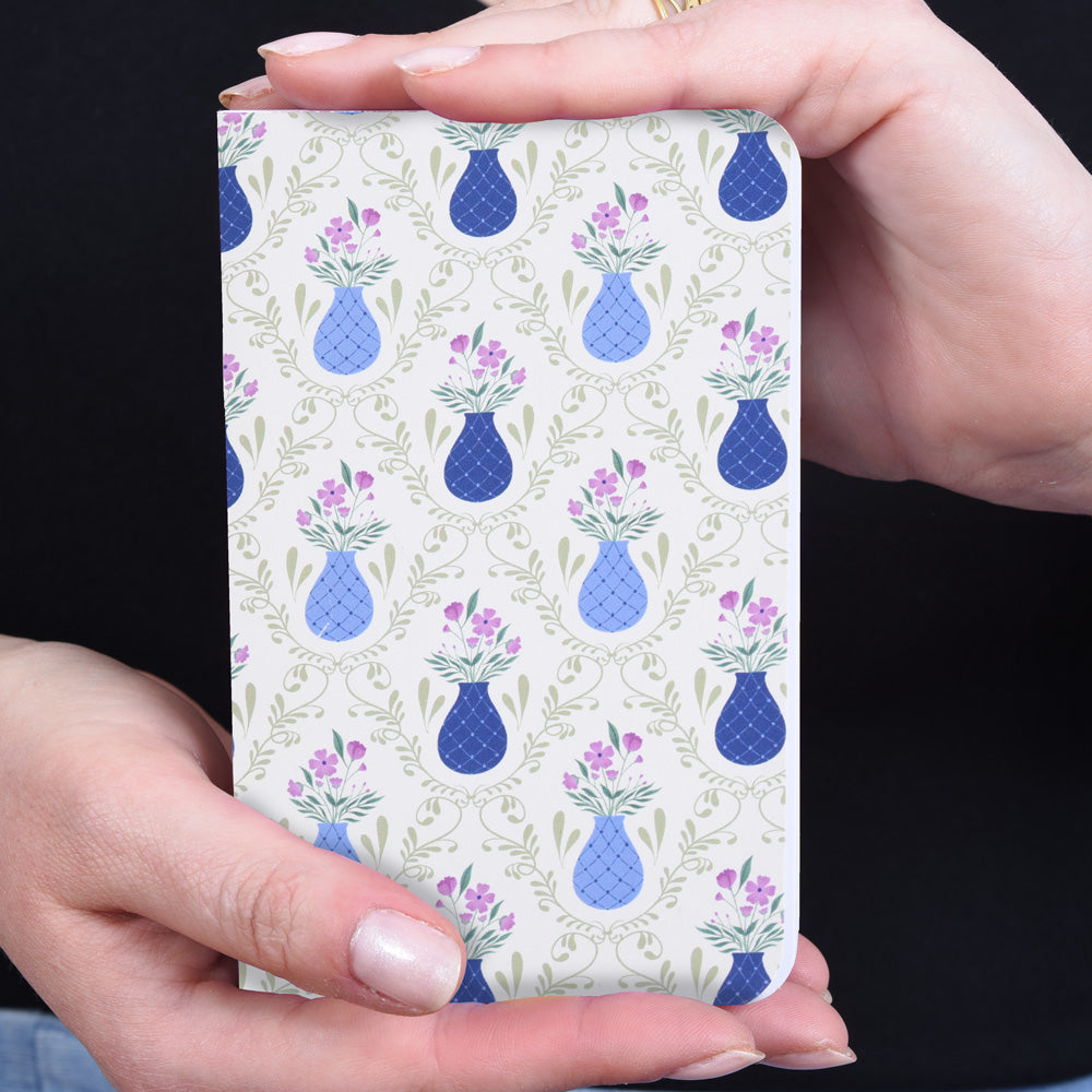 hands holding a small pocket notebook with a pattern of blue vases and purple flowers on a sage green background