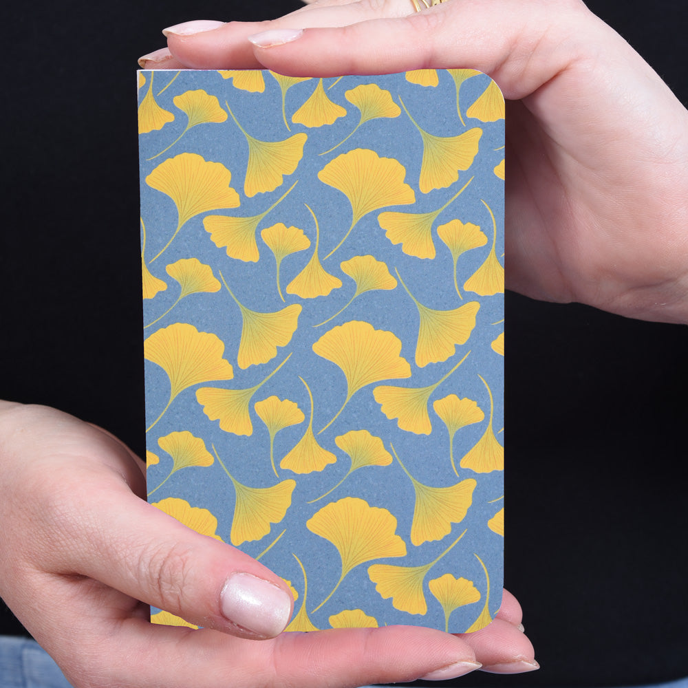 hands holding a small pocket notebook with a pattern of yellow ginkgo leaves on a blue background