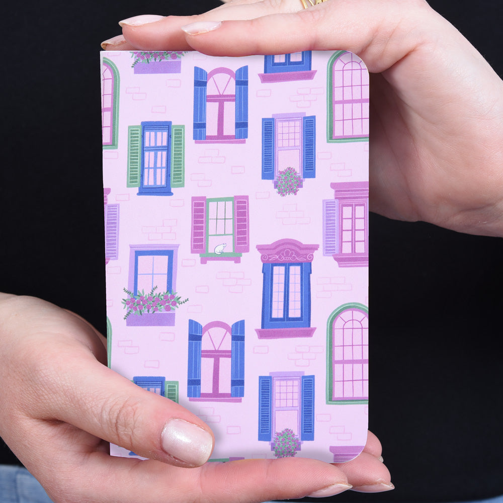 hands holding a small pocket notebook with a pattern of windows and flower boxes on a pink brick background