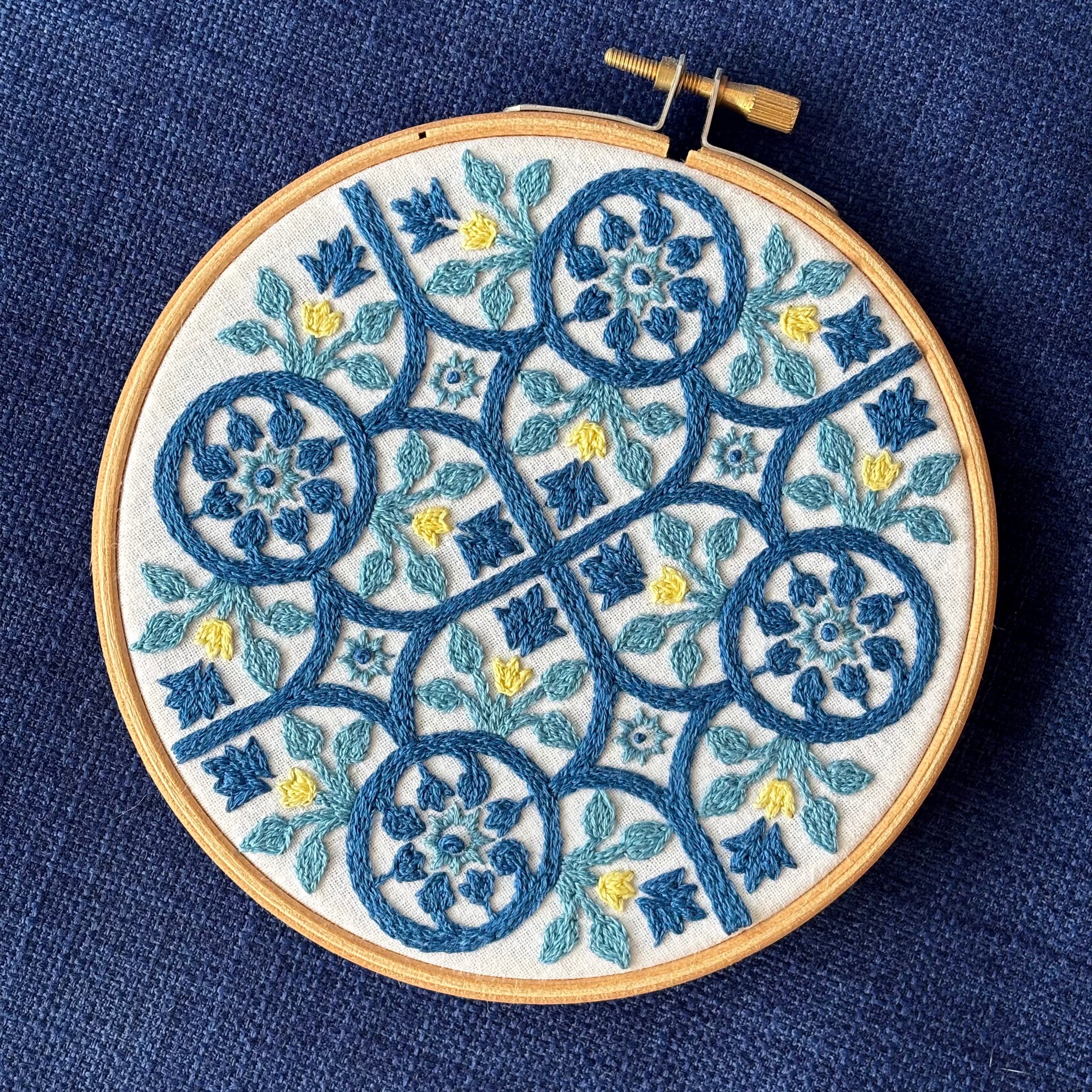 Dark blue, light blue, and yellow embroidery based on traditional Portuguese tile with flowers, leaves, lines, and stars. Fabric is white on white cotton. Embroidery is displayed as wall art in light wood embroidery hoop.