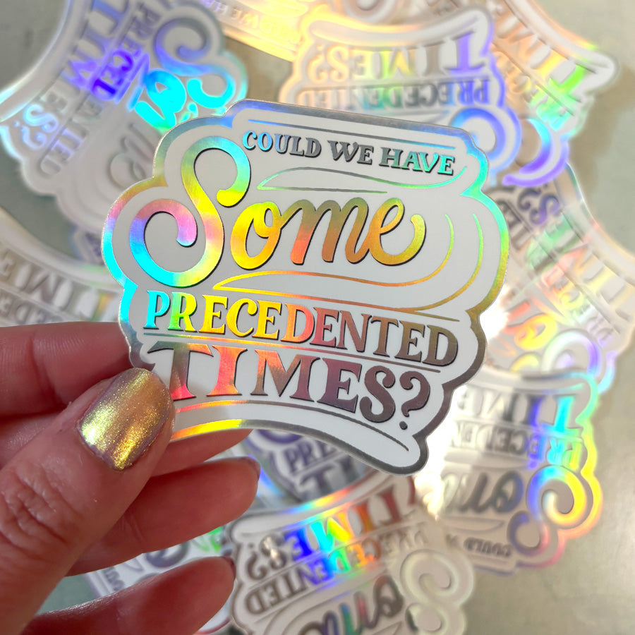 Holographic sticker with colorful text that says "could we have some precedented times?" held by a hand