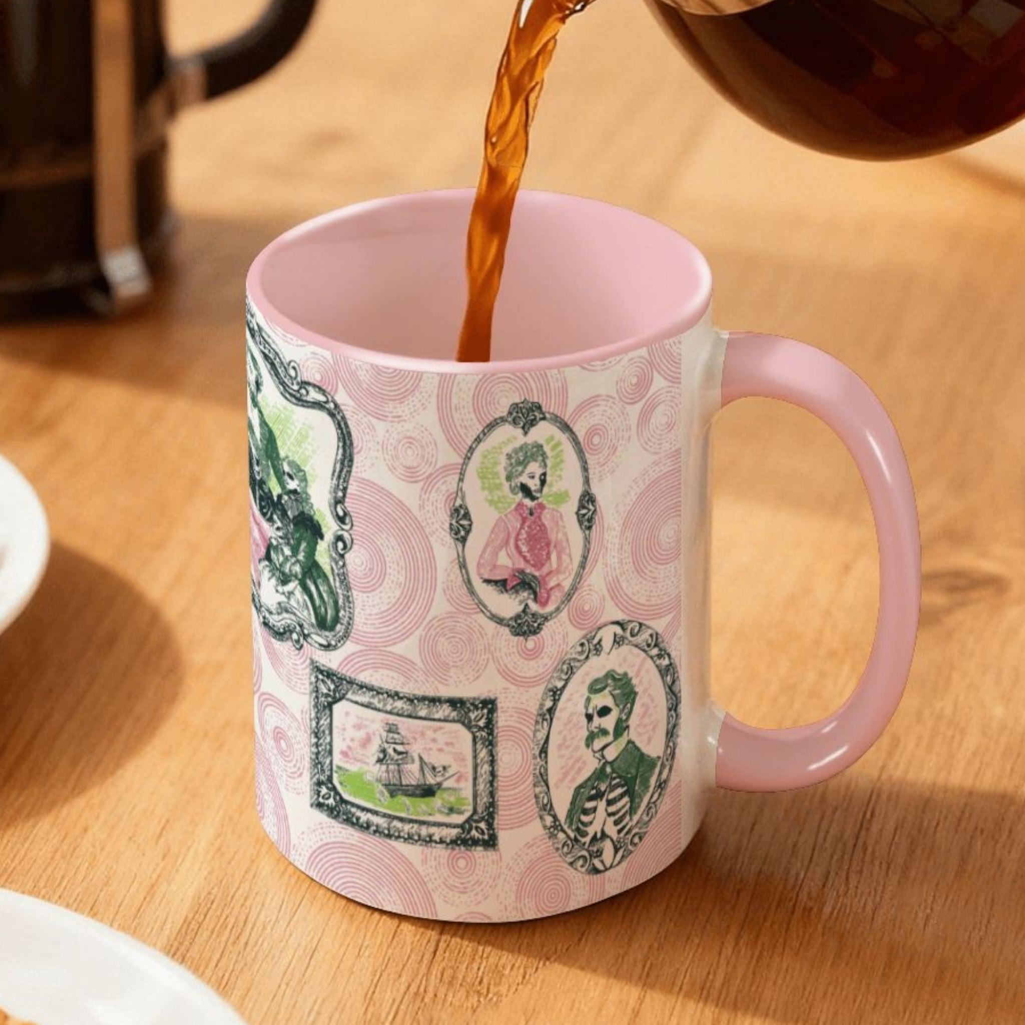Coffee pouring into a pink-handled preppy portrait mug on a wood table, pink and green skull-faced gallery frames