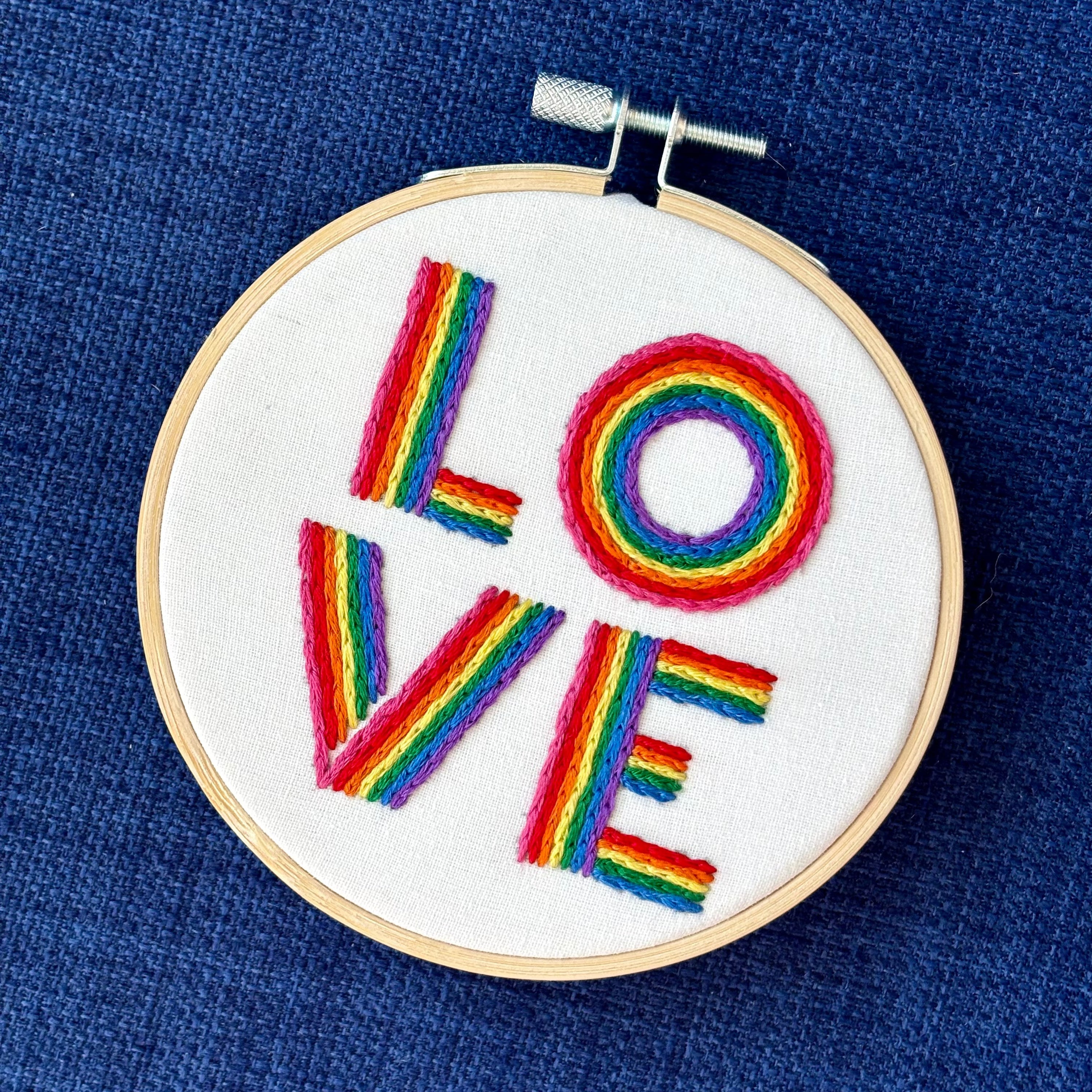 The word "love" is hand embroidered on two lines, in a square, in all capital letters. Each letter is stitched in stripes of rainbow colors: pink, red, orange, yellow, green, blue, purple. Text is sewn on white cotton. Framed as wall art in light wood embroidery hoop.
